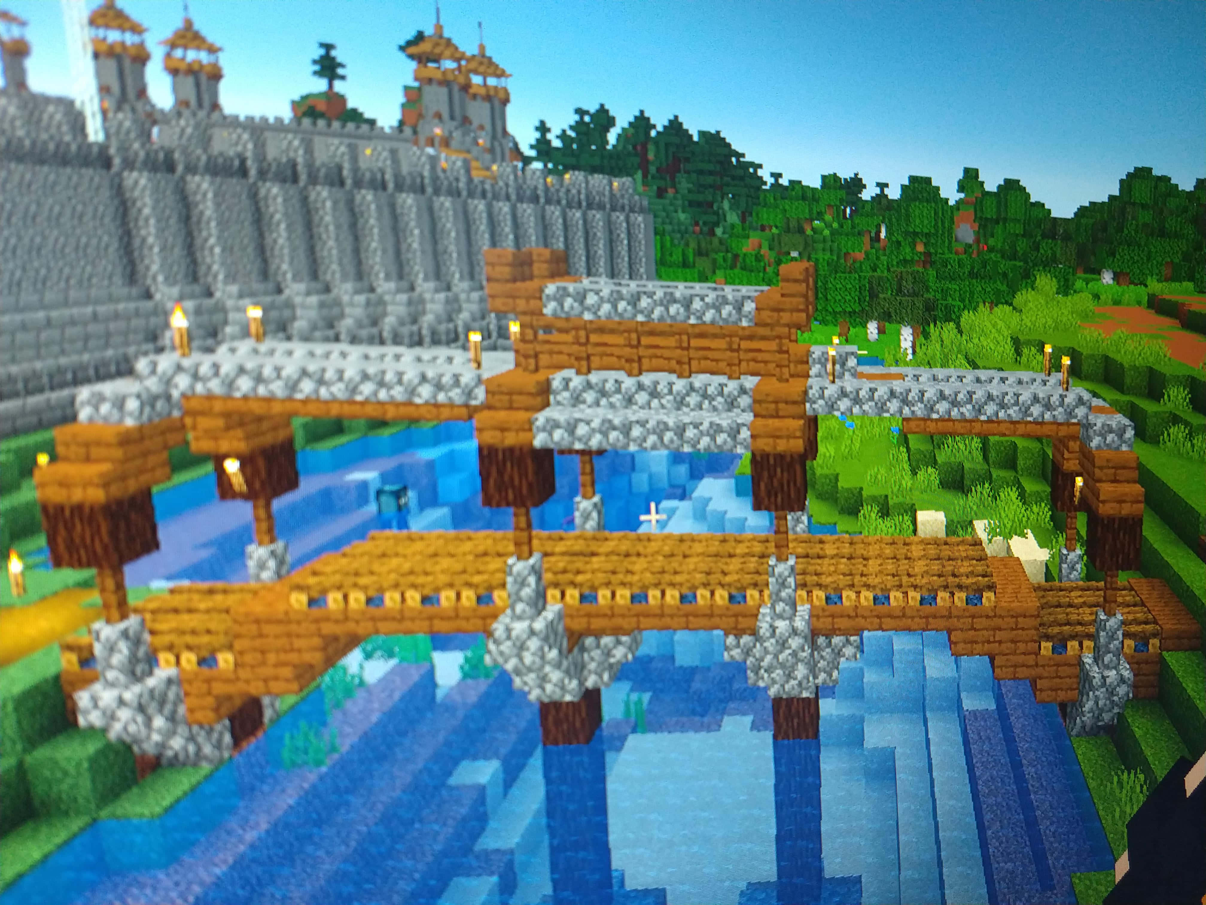 Minecraft Bridges 桌布 壁紙 — 一條像素化的Minecraft橋樑，帶有火炬，橫跨藍色河流，通往被綠樹環繞的石頭城堡。