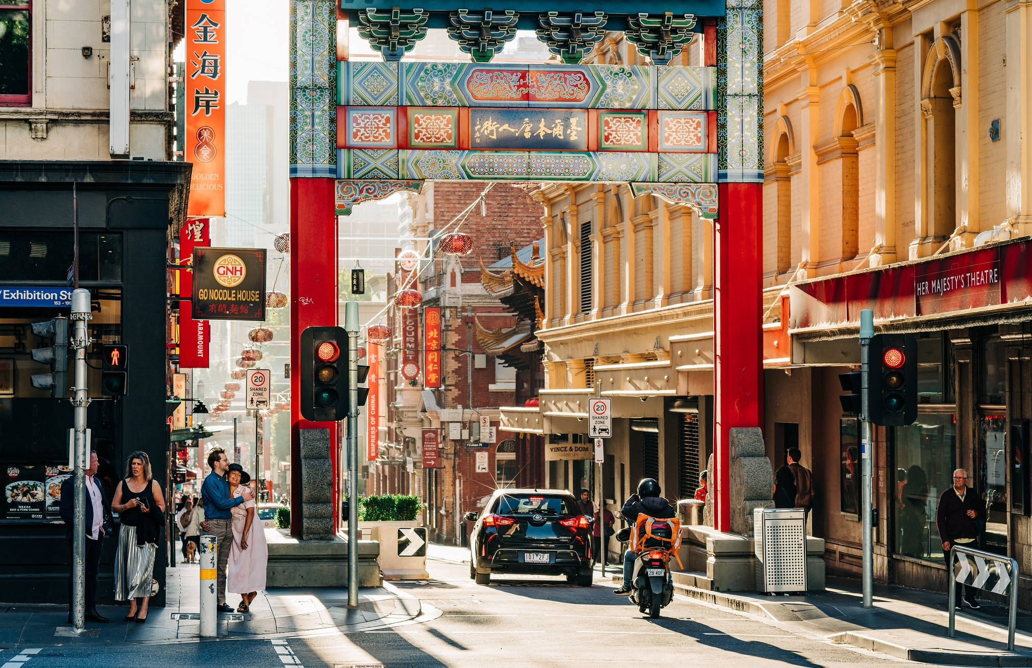 Melbourne 桌布 壁紙 — 一條充滿活力的城市街道，以色彩繽紛的傳統中式牌坊為框架，行人與車輛在明亮的日光下穿梭。
