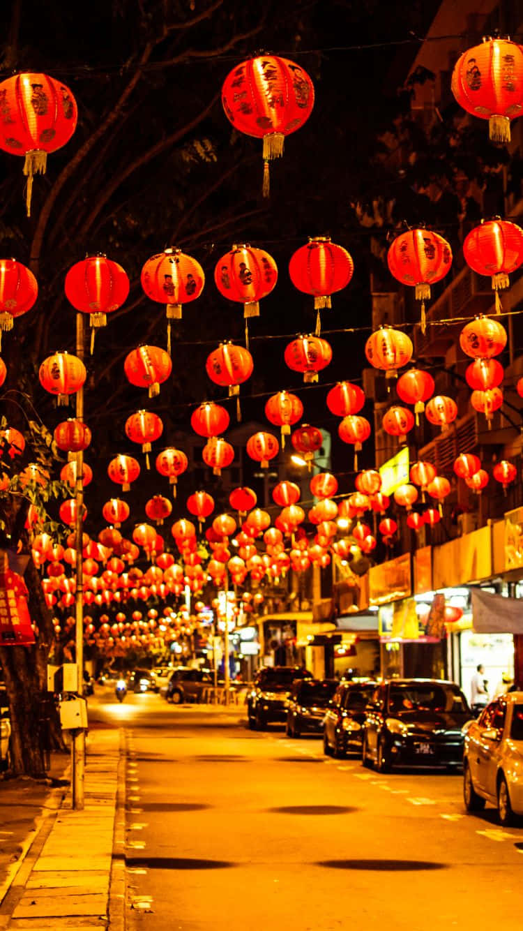 New Year 桌布 壁紙 — 一條充滿活力的夜晚街道，掛滿了成排的紅色發光燈籠，懸掛在停泊的汽車和明亮的商店櫥窗上方。