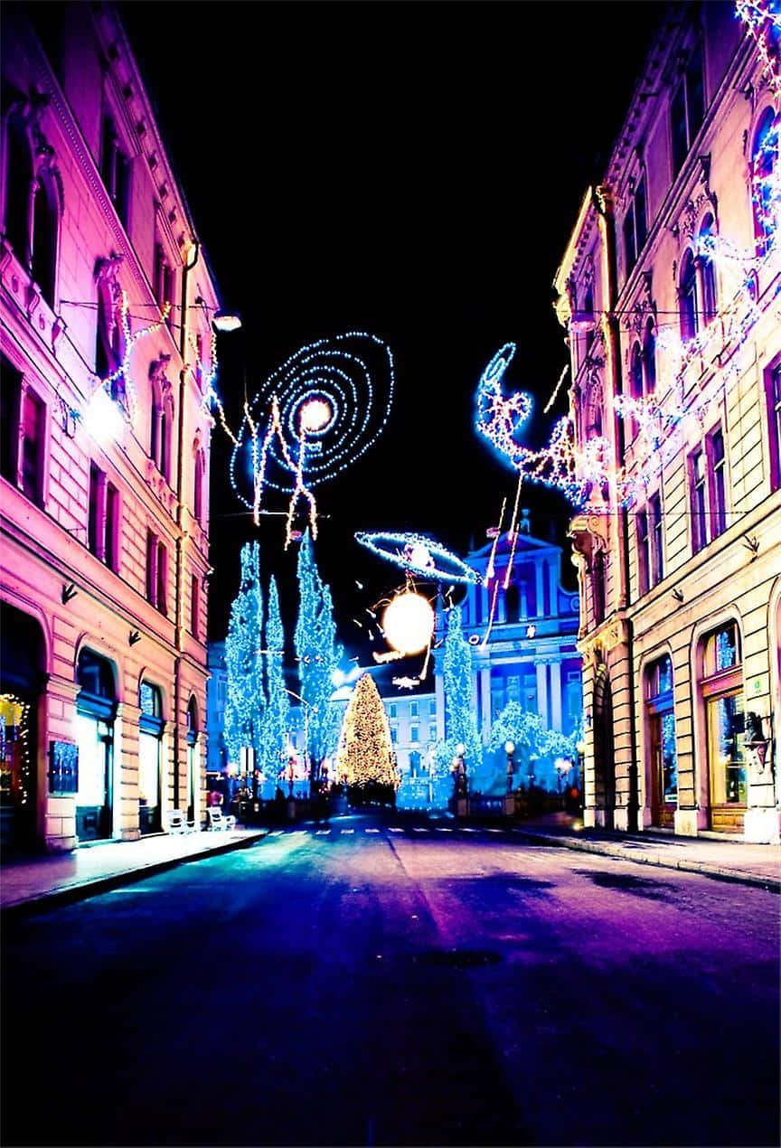 Christmas Eve 桌布 壁紙 — 一條充滿活力的夜間城市街道，裝飾著耀眼的藍色和紫色節日燈飾，盡頭有一棵閃閃發光的聖誕樹。