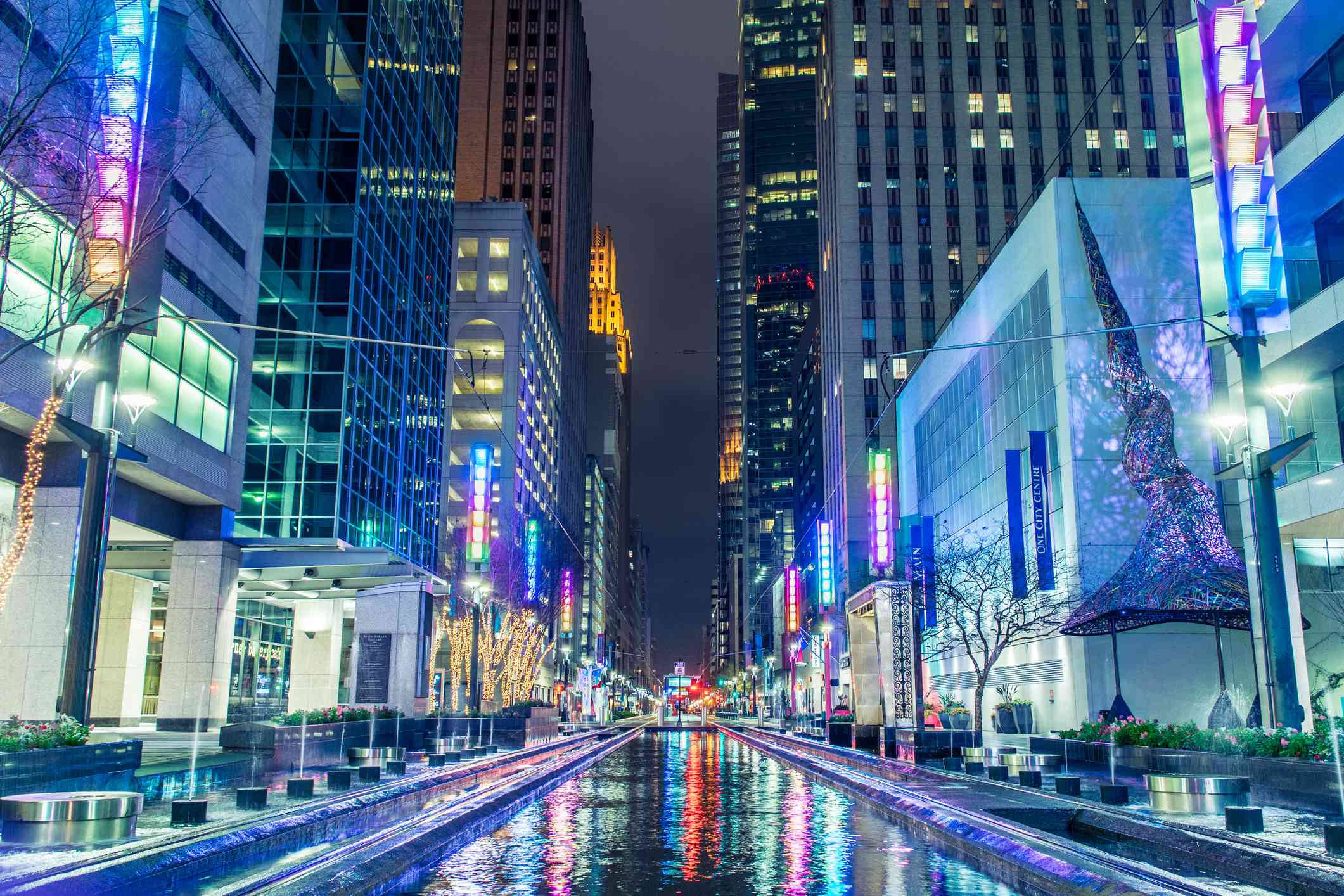 Houston 桌布 壁紙 — 一條充滿活力的夜間城市街道，高樓大廈閃爍著色彩繽紛的霓虹燈，倒映在濕漉漉的中央運河中。