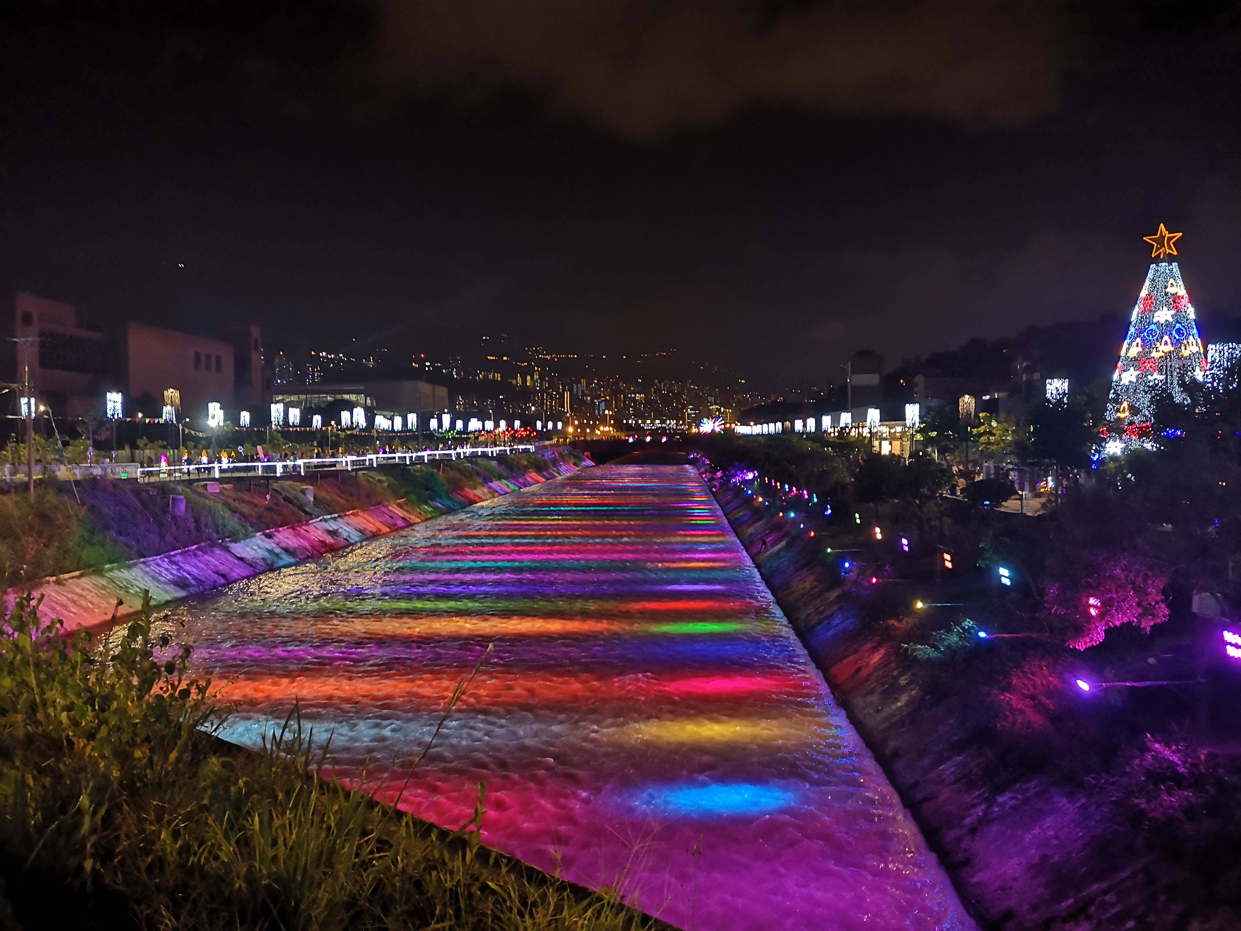 Medellin 桌布 壁紙 — 一條充滿活力的夜間河流，被彩虹燈光照亮，流經一個節日氣氛濃厚的城市公園，公園內有一棵發光的聖誕樹。