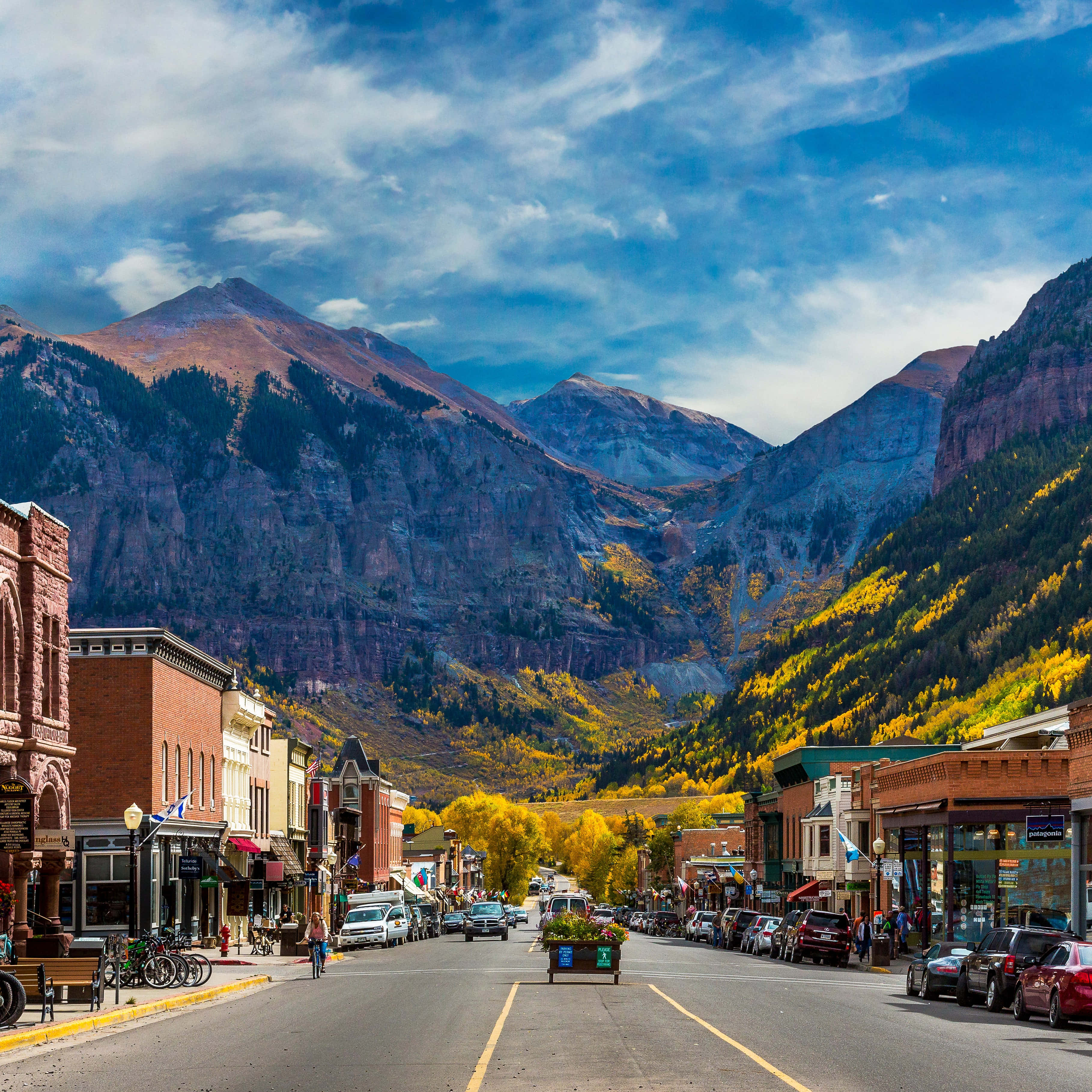 Colorado Mountain 桌布 壁紙 — 一條充滿活力的山城街道，兩旁是磚砌建築和秋樹，上方是明亮的藍天，點綴著零星雲朵。