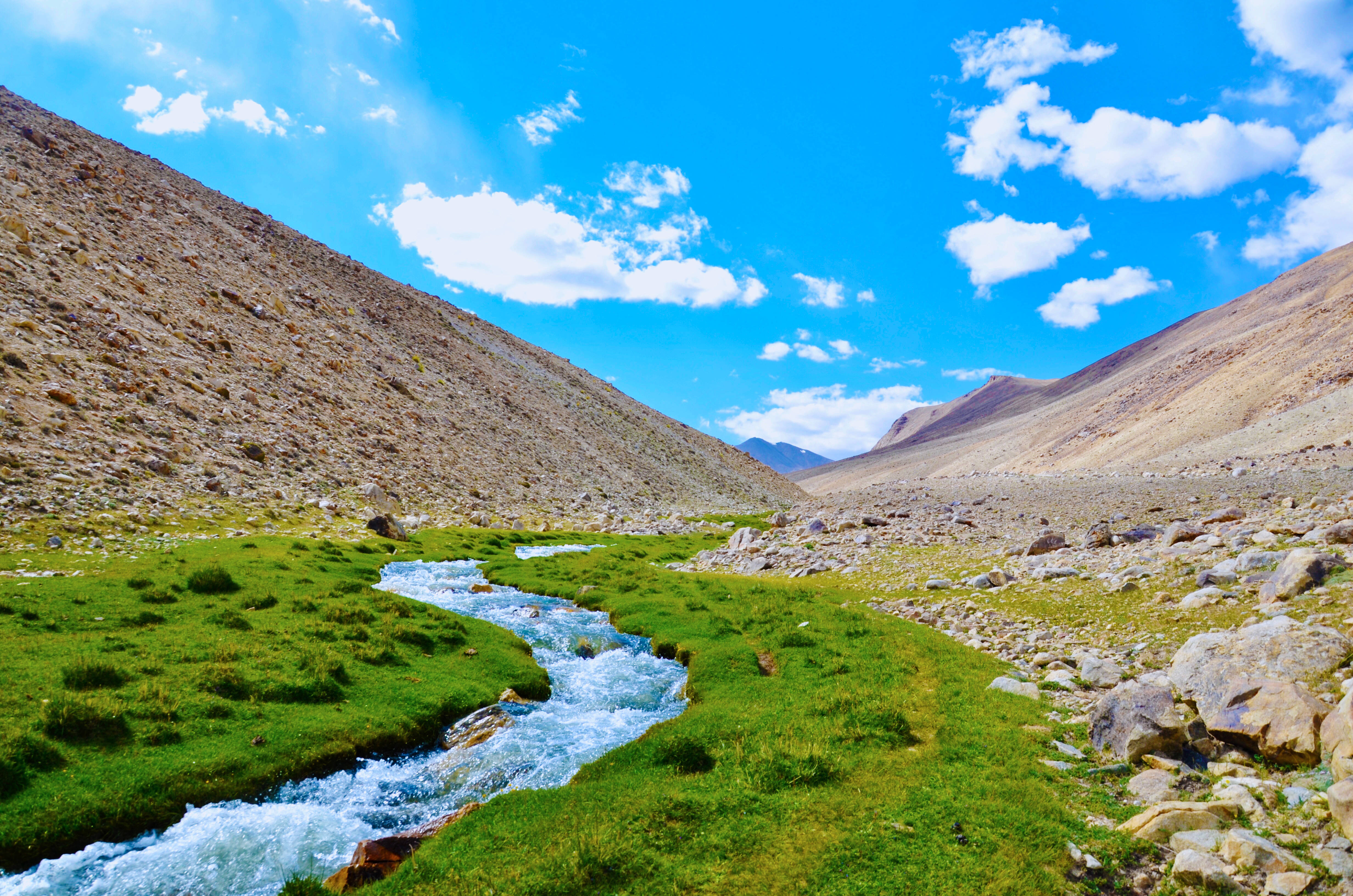 Tajikistan 桌布 壁紙 — 一條充滿活力的山間溪流蜿蜒穿過鬱鬱蔥蔥的綠色草地，上方是明亮的藍天，點綴著蓬鬆的白雲。