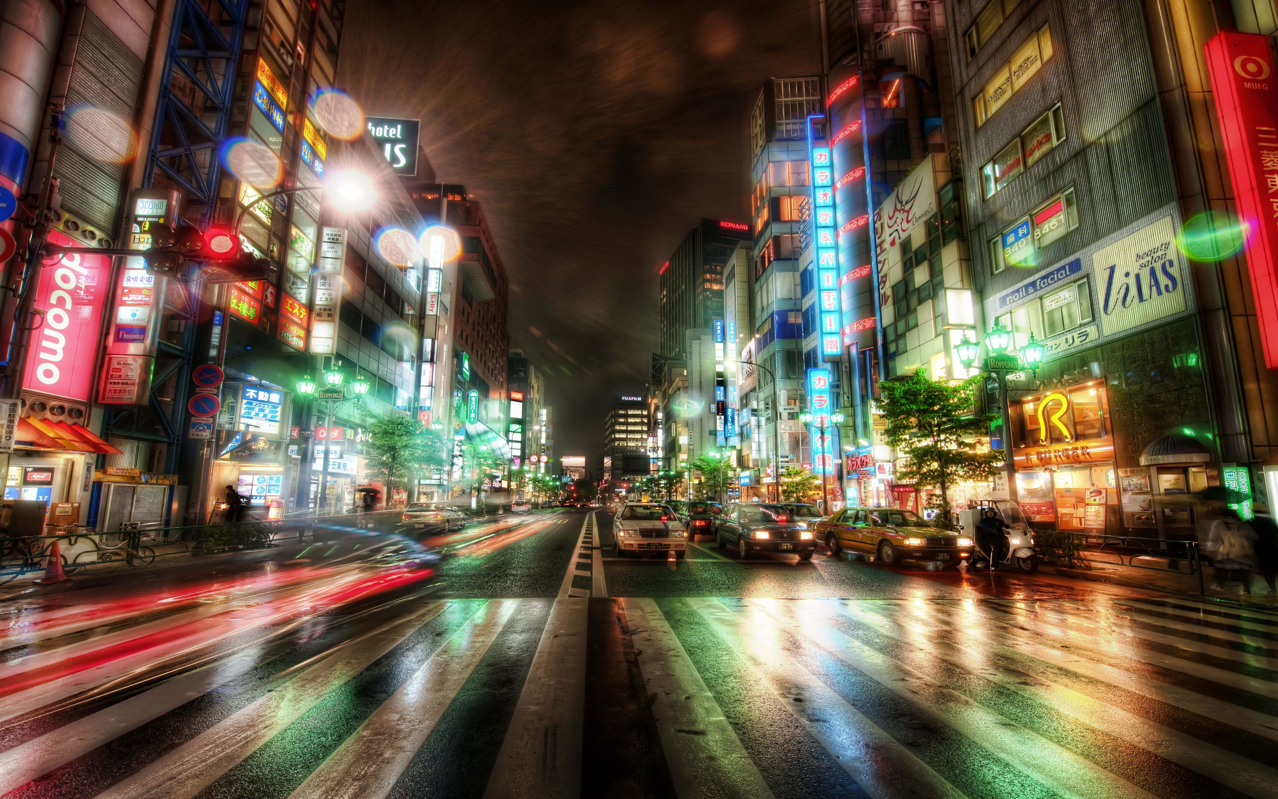Buildings 桌布 壁紙 — 一條充滿活力的東京街道，夜晚霓虹燈閃爍，車燈在濕漉漉的路面上留下流動的光影。