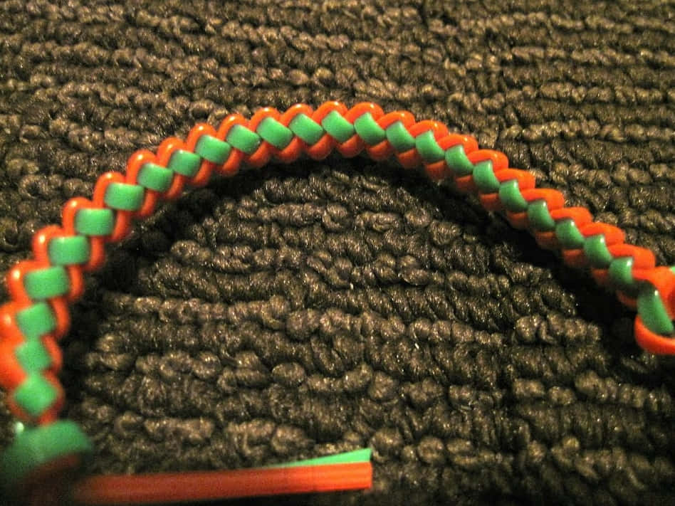 Butterfly Stitch 桌布 壁紙 — 一條充滿活力的橙色和綠色編織手鐲放在深色紋理布料表面上。