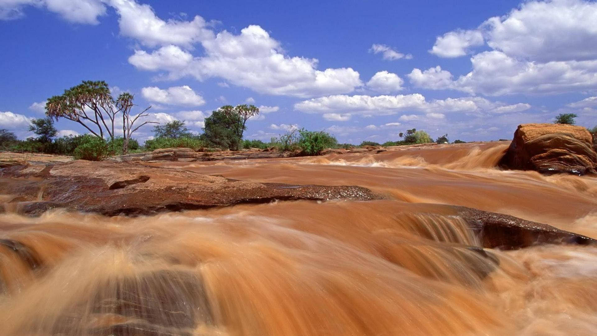 Kenya 桌布 壁紙 — 一條充滿活力的橙色河流在岩石地形上奔流，上方是點綴著蓬鬆白雲的明亮藍天。