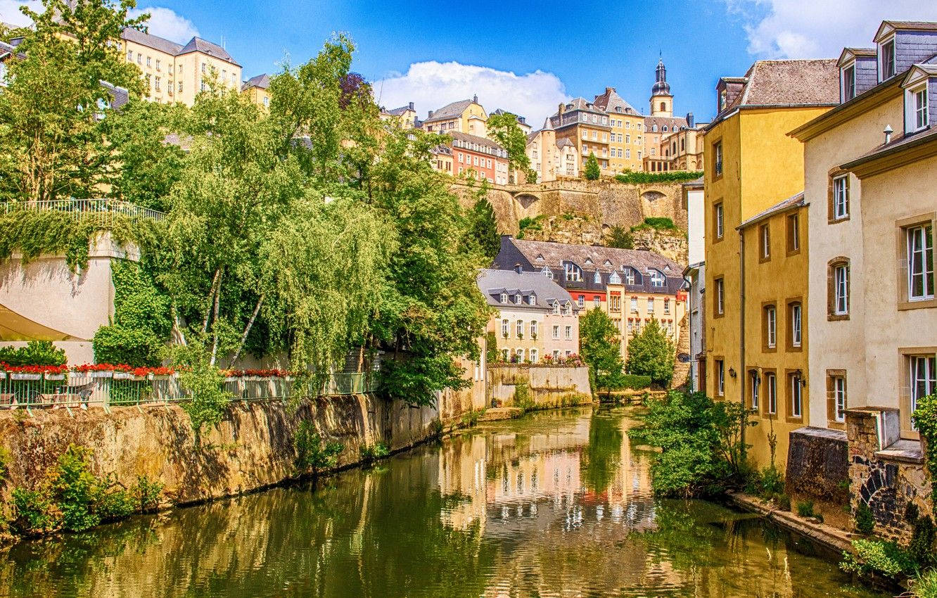 Luxembourg 桌布 壁紙 — 一條充滿活力的歐洲運河蜿蜒穿過歷史悠久的石頭建築，在明亮的藍天下有蓬鬆的雲朵。