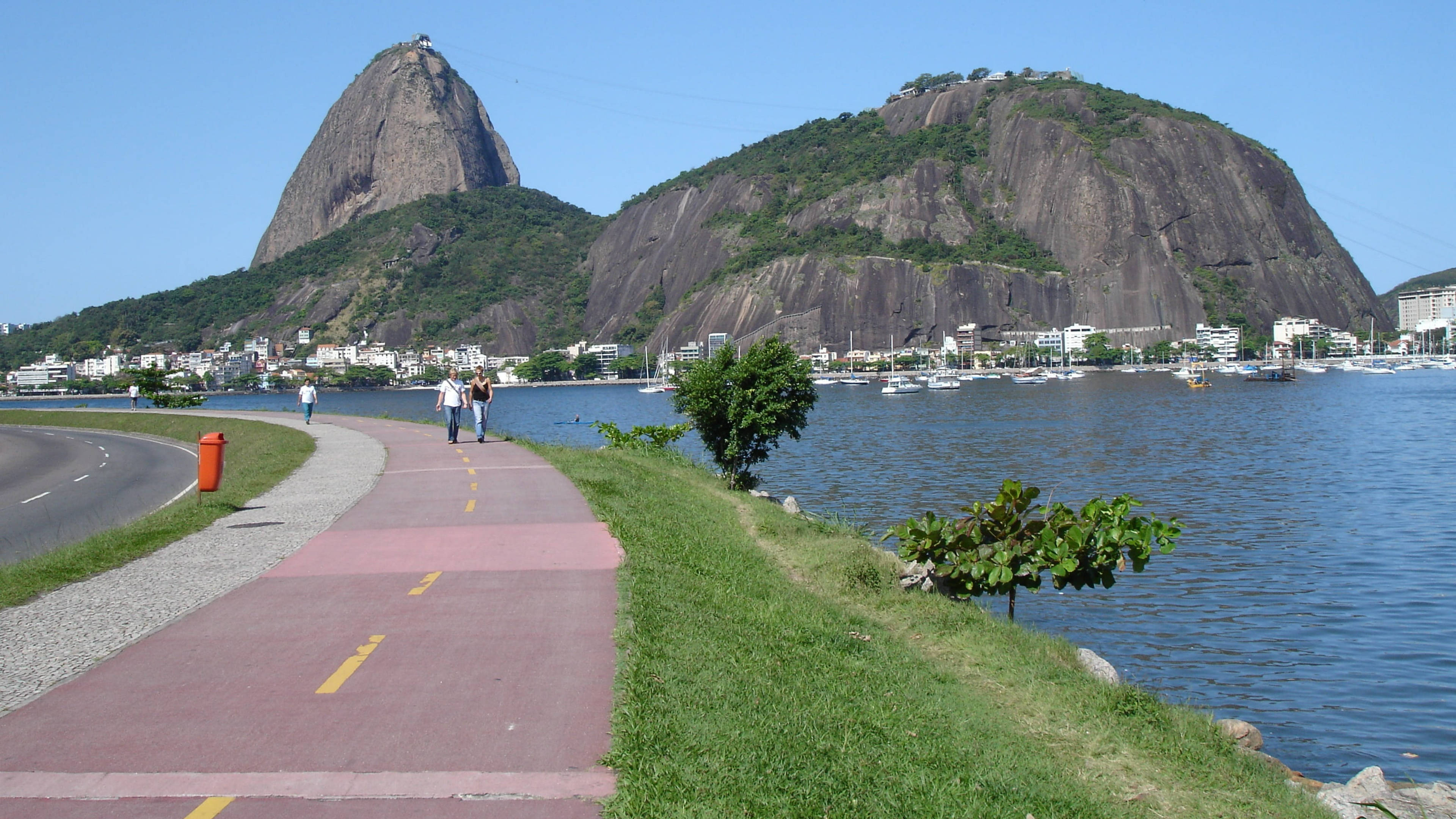 rio 桌布 壁紙 — 一條充滿活力的紅色小徑蜿蜒在草地岸邊，旁邊是平靜的藍色海水，標誌性的糖麵包山在清澈的藍天下聳立在背景中。