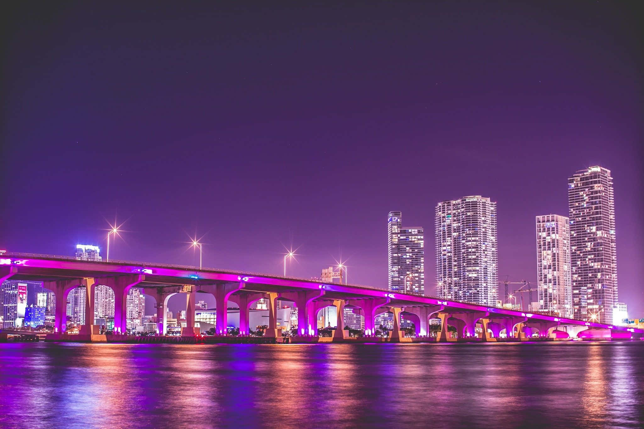 桌布 壁紙 — 一條充滿活力的紫色燈光橋樑橫跨平靜的河流，夜晚中發光的摩天大樓倒映在水面上。