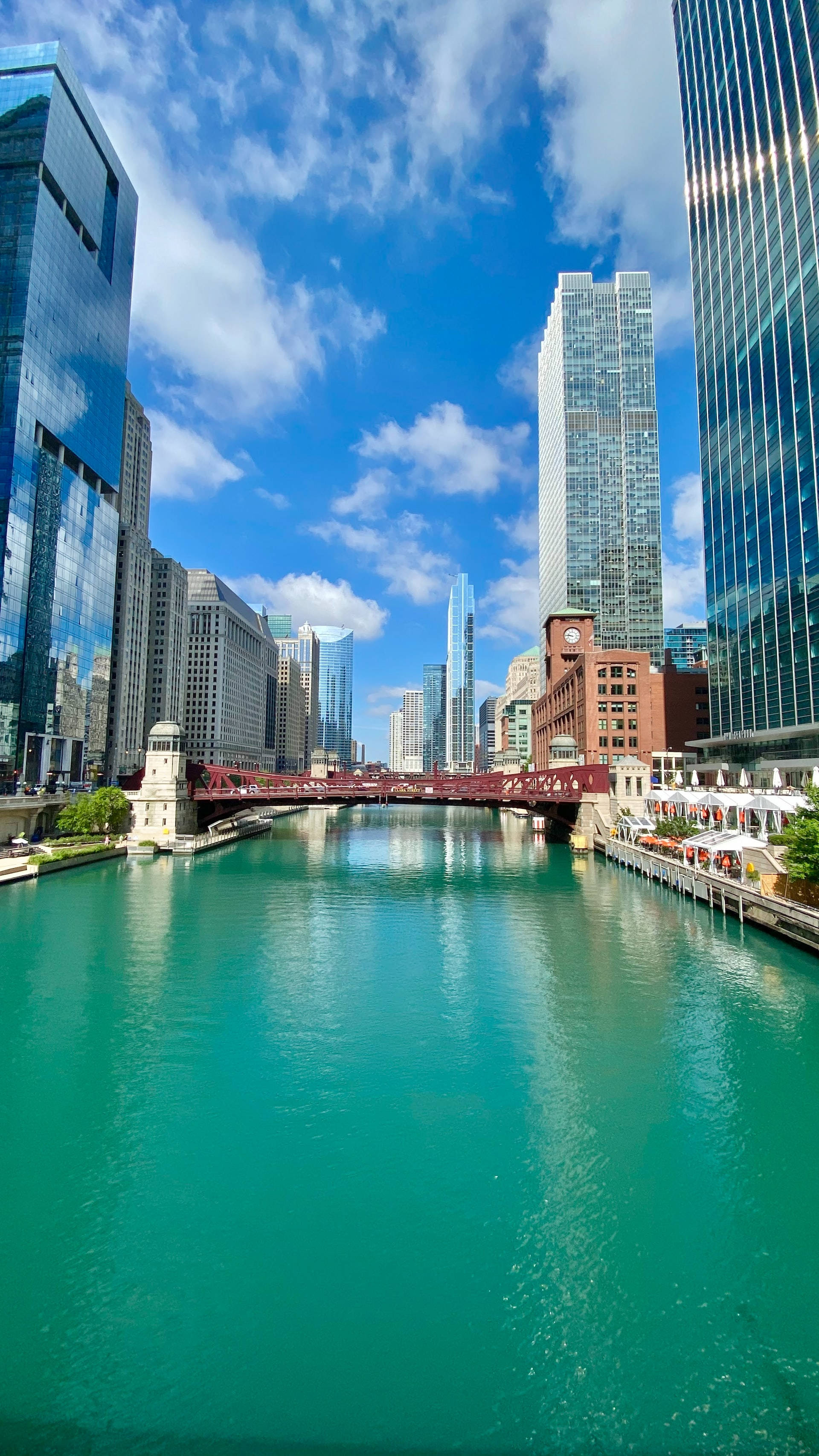 Skyscraper 桌布 壁紙 — 一條充滿活力的藍綠色河流流經現代城市景觀，有高聳的玻璃摩天大樓和一座紅色橋樑，在明亮的藍天下有蓬鬆的雲朵。