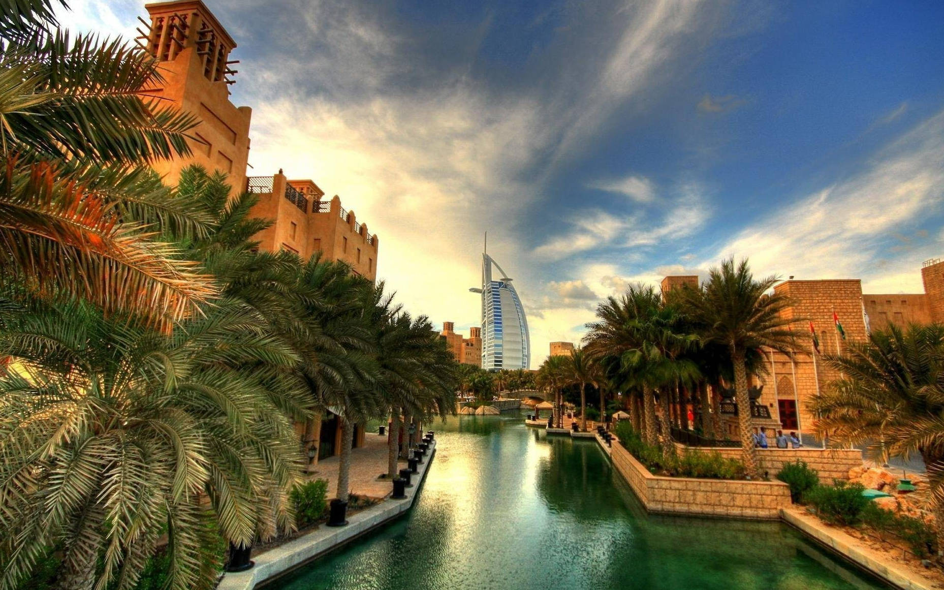 Arab 桌布 壁紙 — 一條充滿活力的運河，兩旁種滿棕櫚樹和傳統建築，遠處的帆船酒店在戲劇性的天空下高聳入雲。