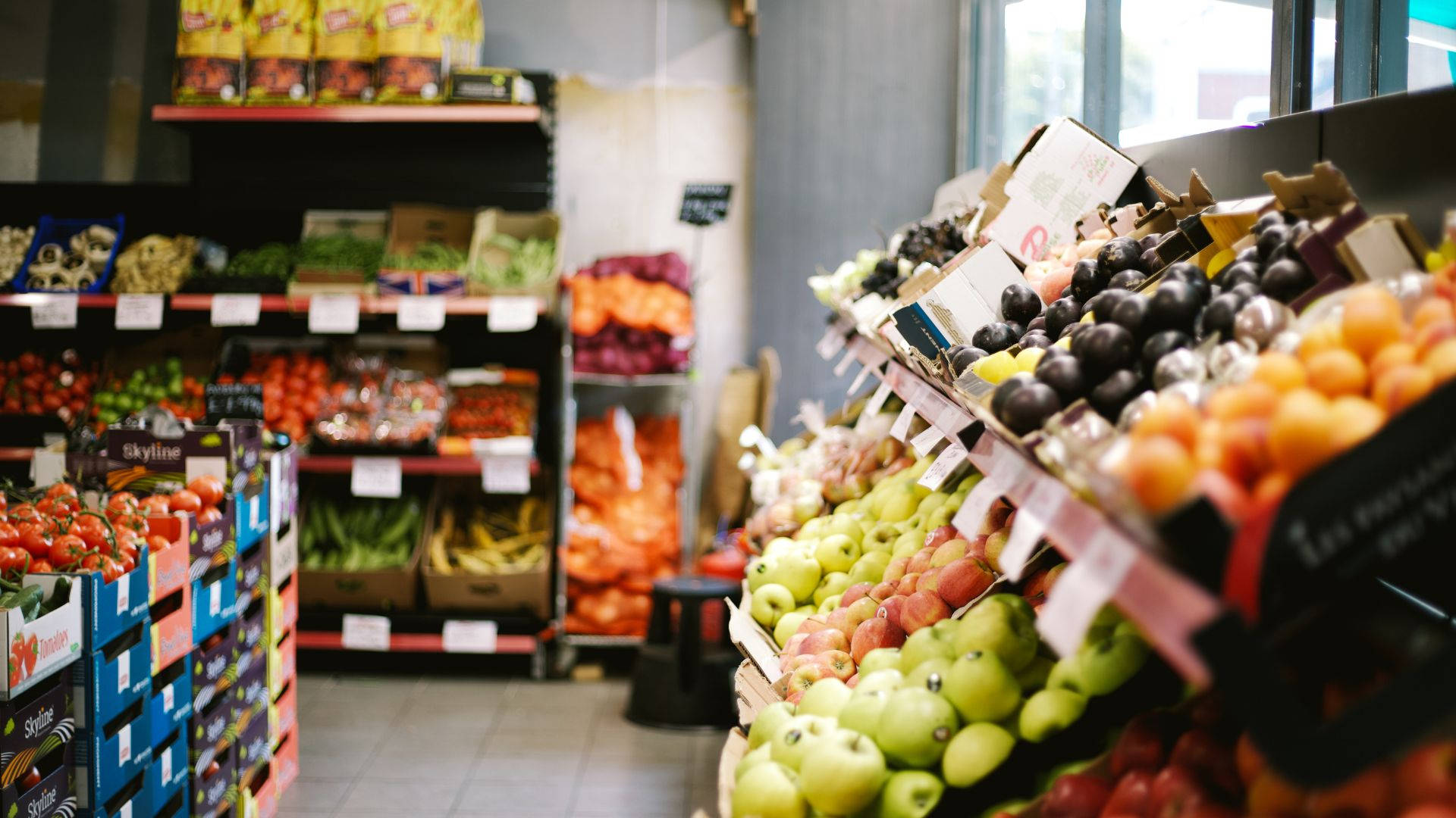 Fruits 桌布 壁紙 — 一條充滿活力的雜貨店通道，擺滿了色彩繽紛的新鮮農產品，如蘋果、李子和番茄，在明亮的燈光下整齊排列。