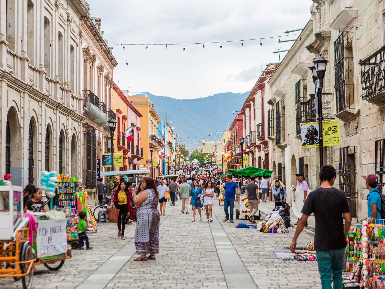 Mexican Food 桌布 壁紙 — 一條充滿活力的鵝卵石街道，位於歷史小鎮，兩旁是色彩繽紛的殖民時期建築，在陰天天空下行人熙熙攘攘。