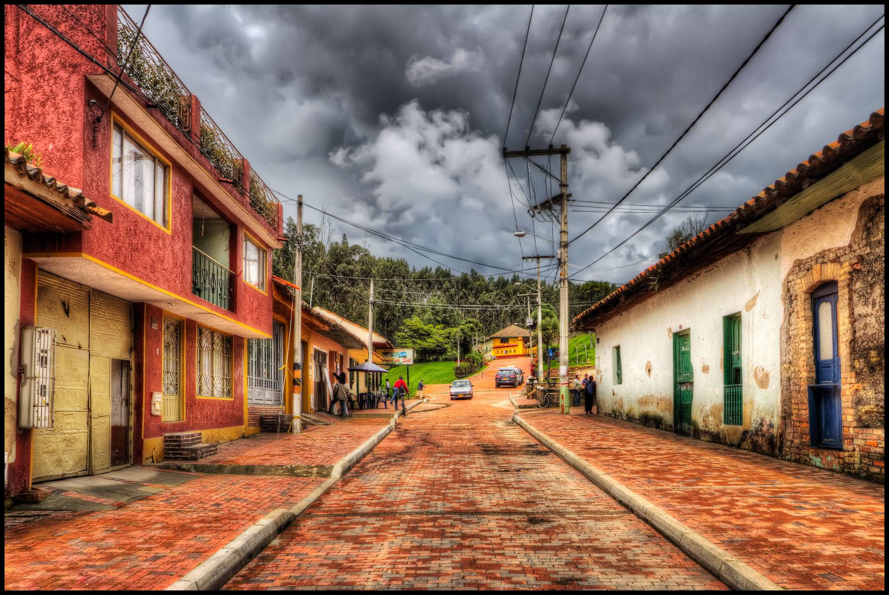 Colombia 桌布 壁紙 — 一條充滿活力的鵝卵石街道，位於色彩繽紛的村莊，天空多雲戲劇性，有行人和汽車。