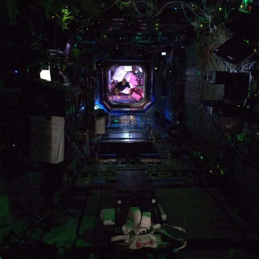 Scary Space 桌布 壁紙 — 一條光線昏暗的太空船走廊，帶有綠色環境照明，通向一個發光的六邊形門口，可以看到穿著粉紅色太空衣的太空人。