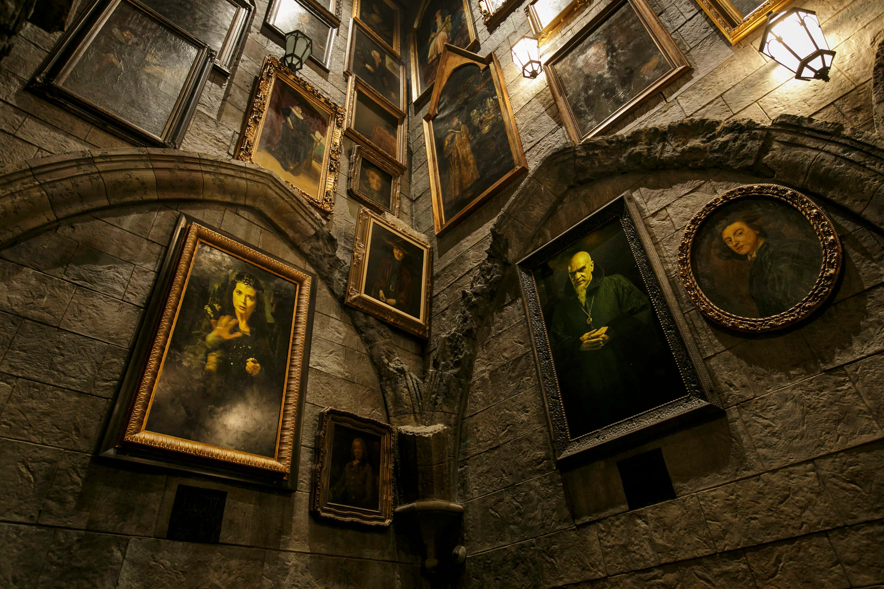 The Hogwarts Dark Arts Classroom 桌布 壁紙 — 一條光線昏暗的石頭走廊，裝飾著眾多在溫暖燈籠光下閃耀的華麗畫像。