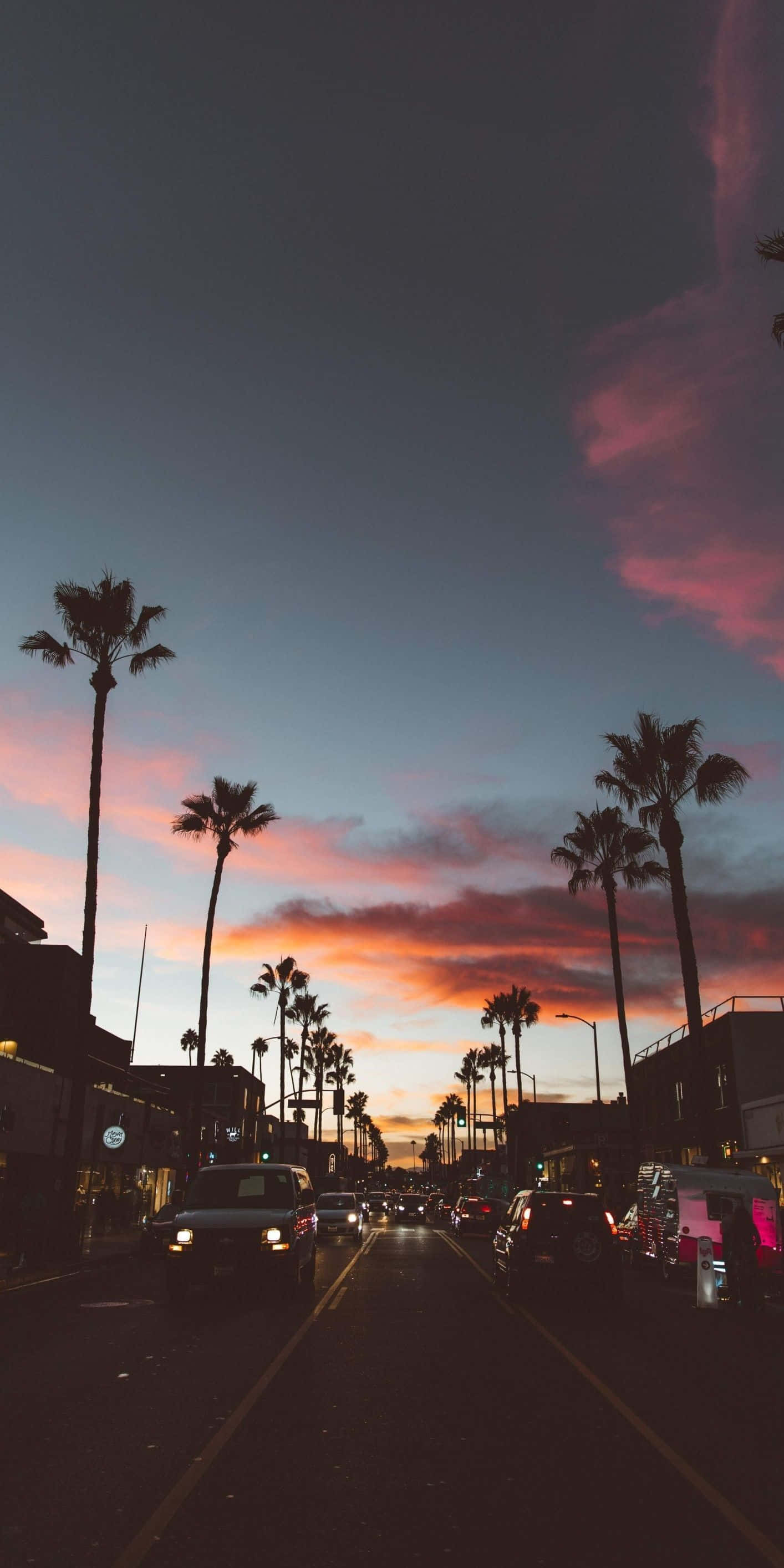 California Aesthetic 桌布 壁紙 — 一條兩旁種滿棕櫚樹的街道，延伸向色彩繽紛的日落天空，呈現粉紅、橙色和紫色調。