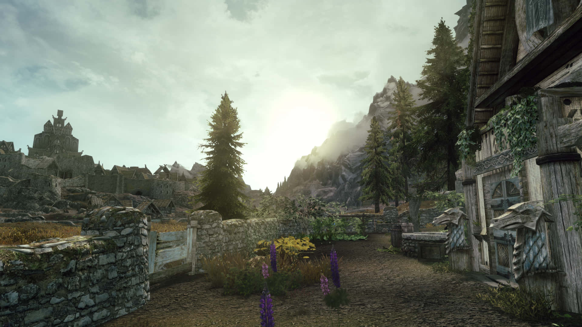 Skyrim Landscape 桌布 壁紙 — 一條古樸的石徑蜿蜒穿過中世紀村莊，村內有木屋和遠處的城堡遺跡，天空多雲。