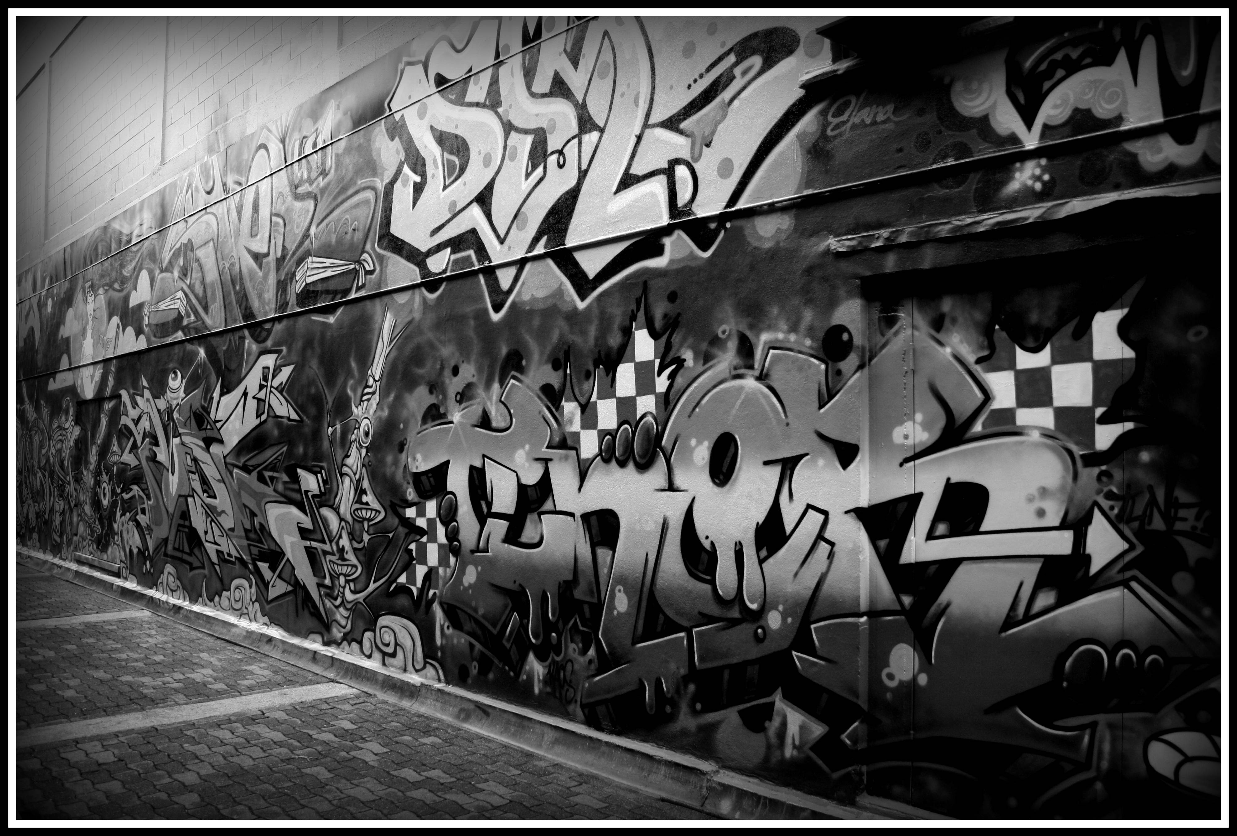 Black And White Graffiti 桌布 壁紙 — 一條單色城市後巷牆壁，上面覆蓋著大膽的塗鴉標籤和帶有棋盤圖案的風格化字體。