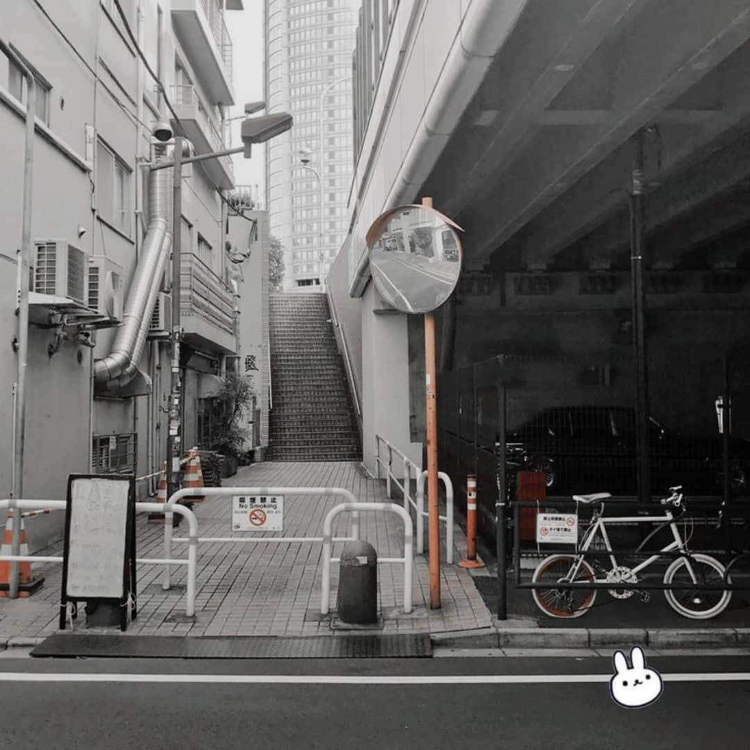 灰色美學動圖 桌布 壁紙 — 一條單色調的城市小巷，有樓梯通往摩天大樓，兩旁是建築物，天橋下停著一輛腳踏車。