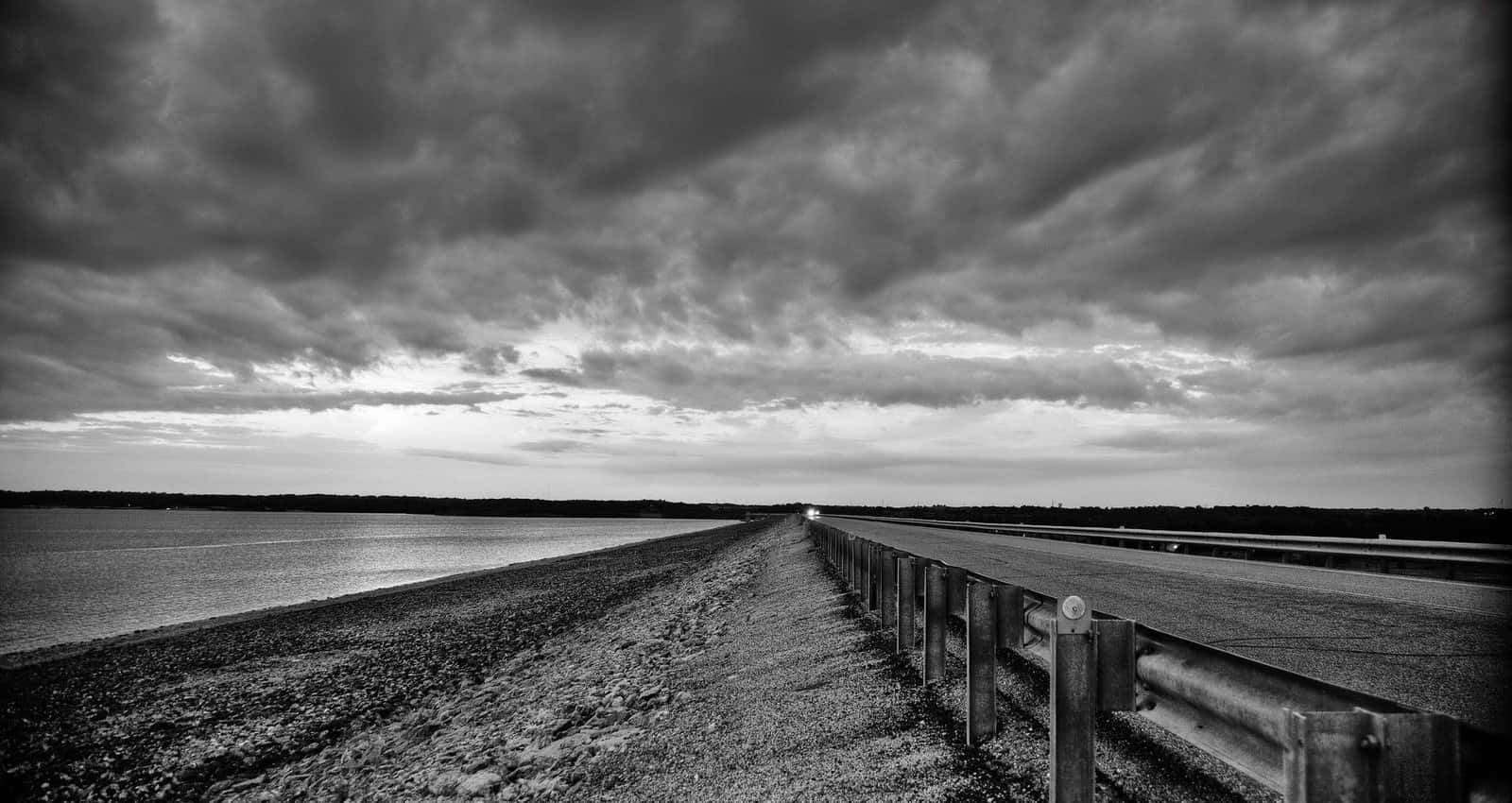 Black And White Sky 桌布 壁紙 — 一條單色道路在戲劇性的多雲天空下，沿著平靜的水體向遠處的汽車延伸。