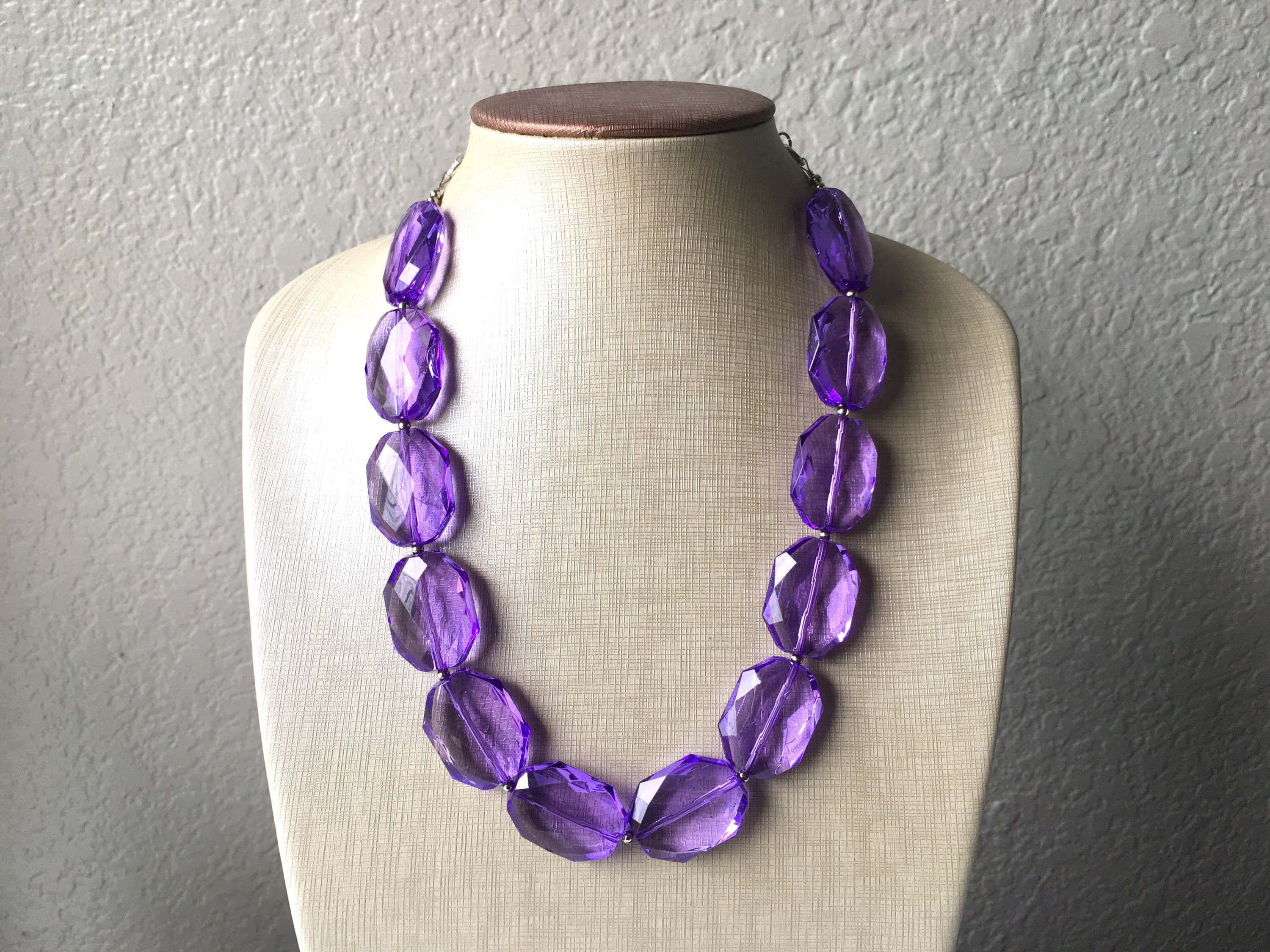 Purple Jewelry 桌布 壁紙 — 一條大膽的紫色刻面珠鏈展示在米色模特兒半身像上，背景是紋理牆壁。