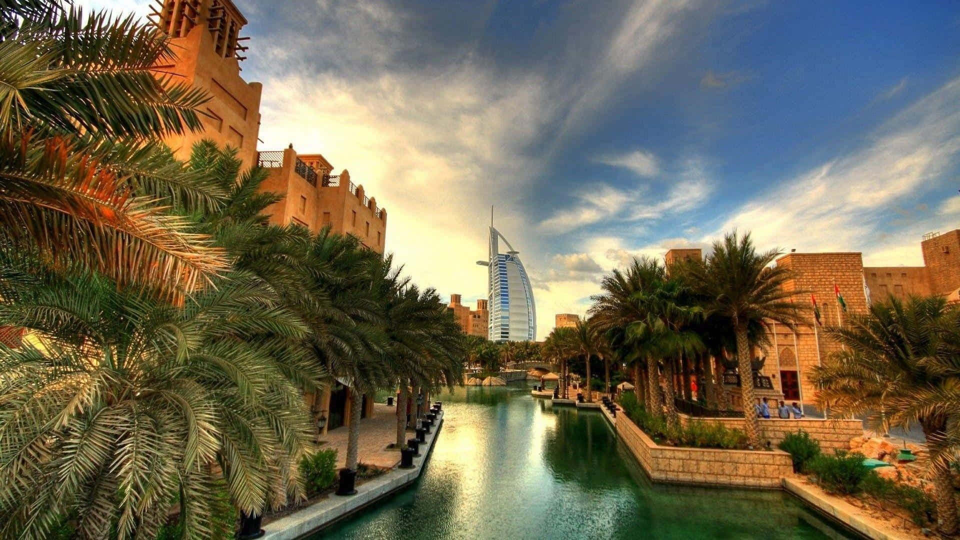 Uae 桌布 壁紙 — 一條大運河，兩旁種滿棕櫚樹和歷史建築，遠處的帆船酒店在戲劇性的天空下高聳入雲。