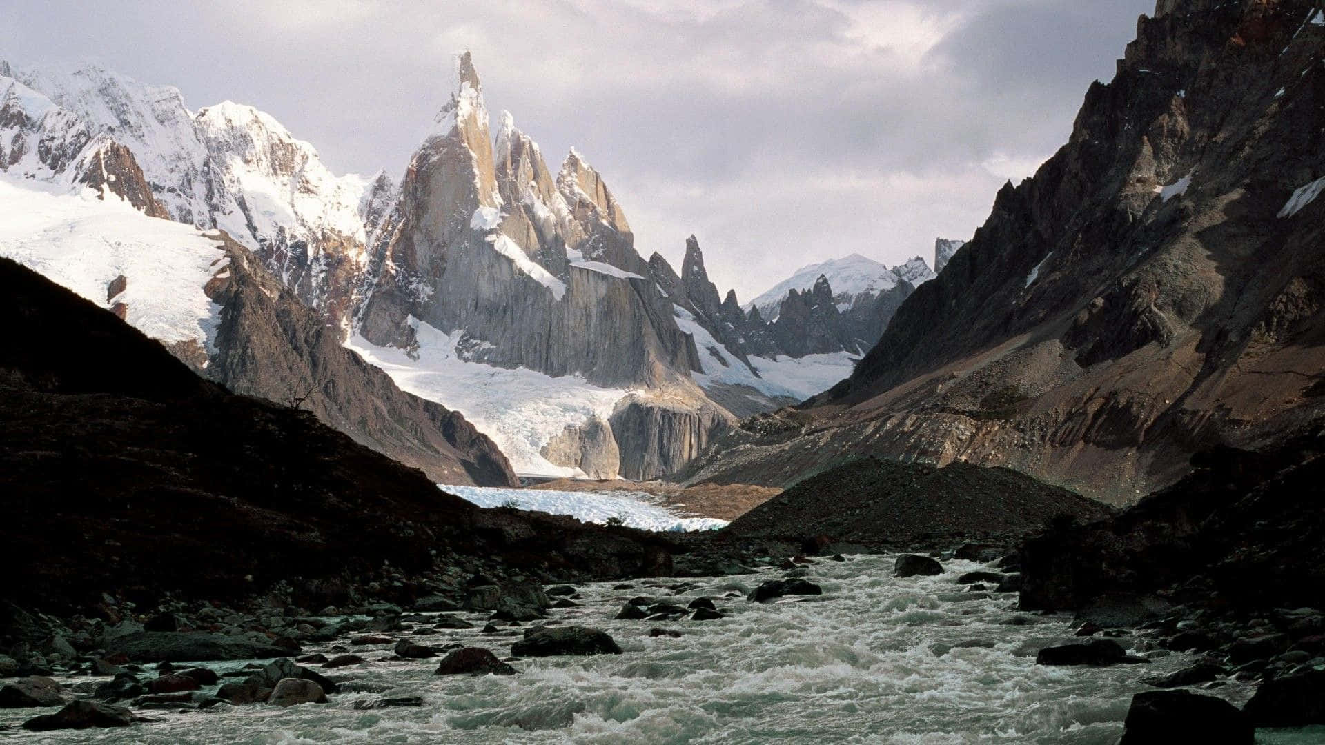 Patagonia 桌布 壁紙 — 一條奔騰的河流在陰天之下，穿過崎嶇的山谷，兩旁是高聳的雪峰。