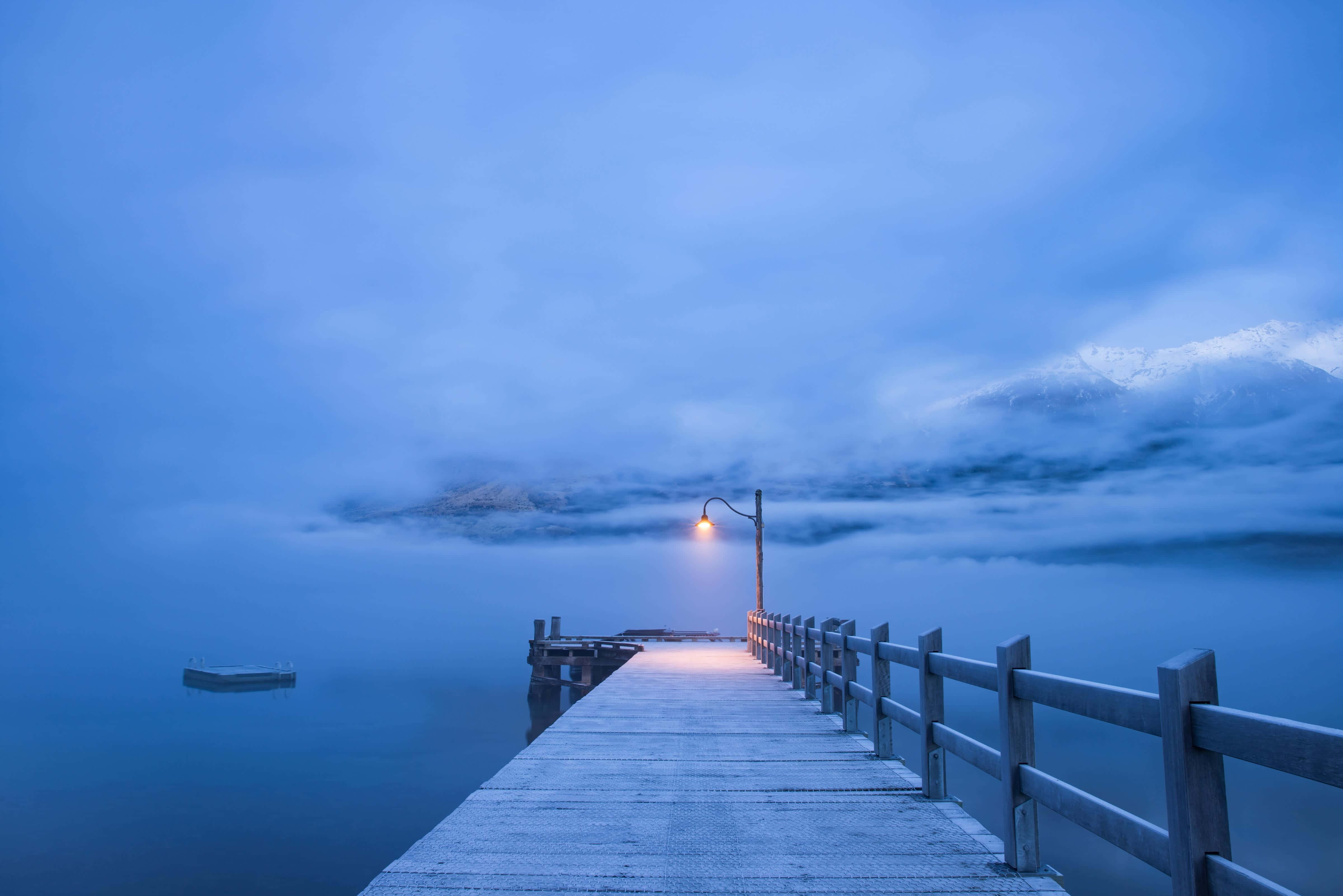 Winter Beach 桌布 壁紙 — 一條孤獨的木碼頭延伸到霧氣瀰漫的湖中，暮色天空下，積雪的山峰在雲層中若隱若現。