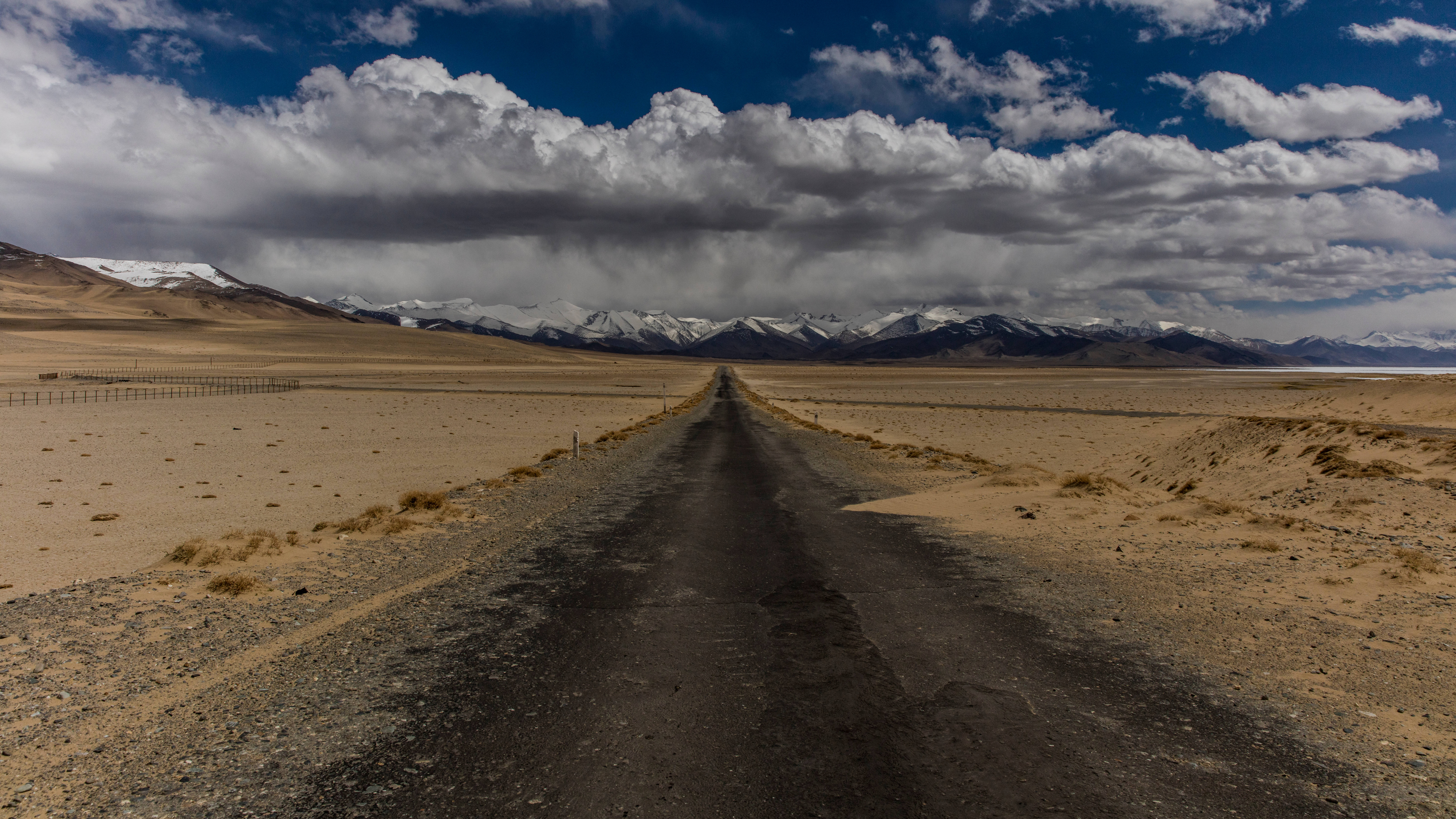 Tajikistan 桌布 壁紙 — 一條孤獨的瀝青公路穿過廣闊的乾旱平原，在戲劇性的多雲天空下延伸至遠方積雪覆蓋的山脈。