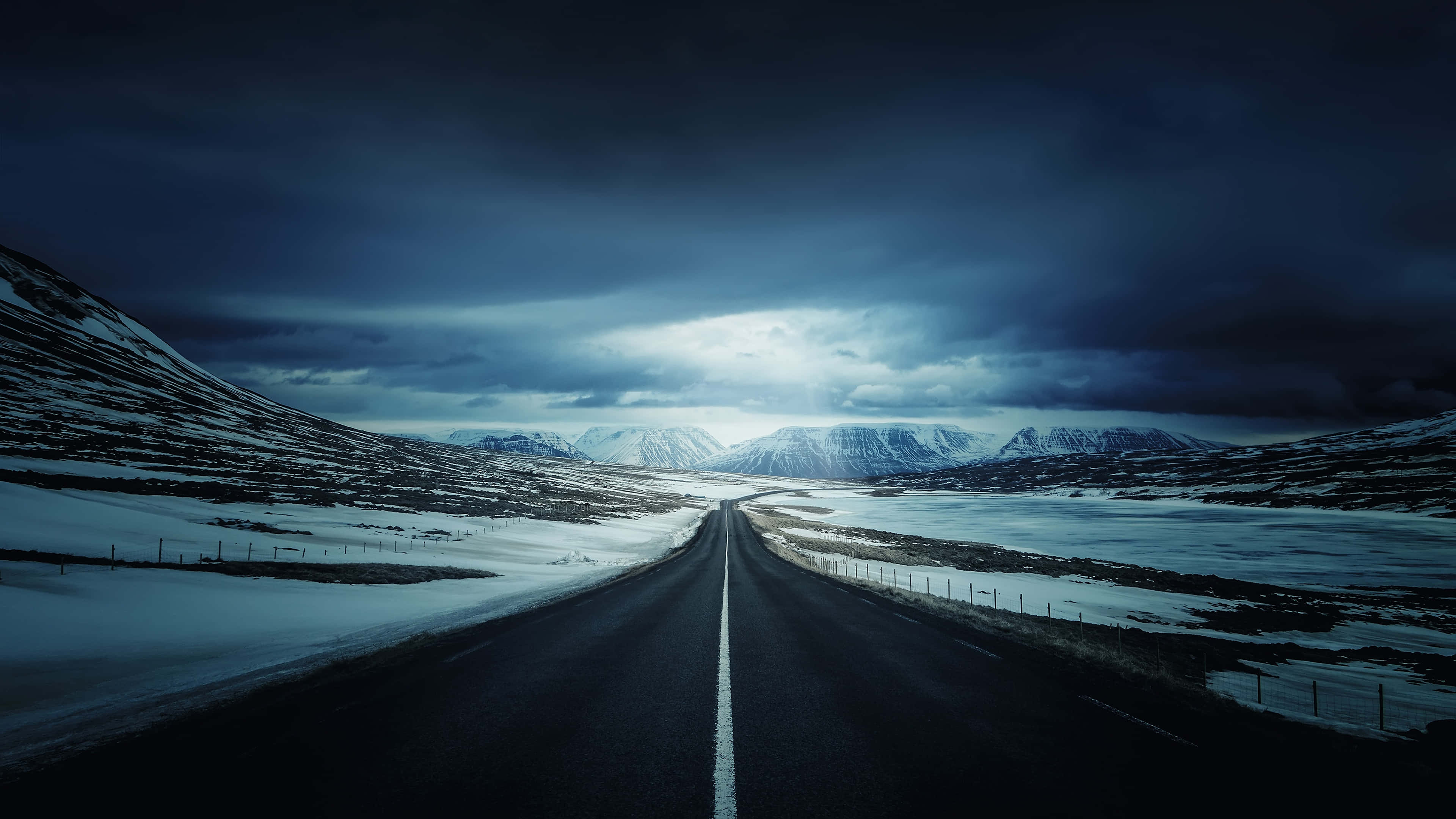 Snowy Road 桌布 壁紙 — 一條孤獨的瀝青路在戲劇性的暴風雨天空下，穿過積雪的山谷，遠處有山峰。