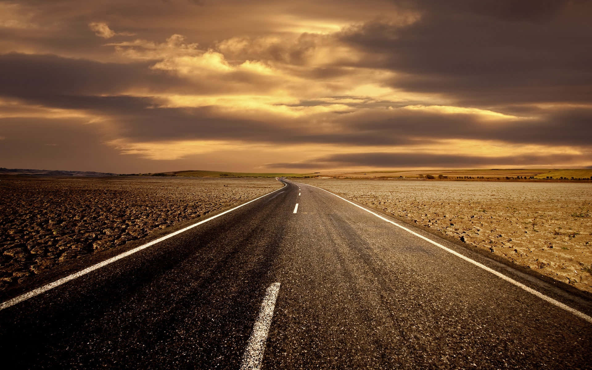 Highways 桌布 壁紙 — 一條孤獨的瀝青路在戲劇性的金色日落天空下，伴隨著厚重的雲層，延伸進廣闊的乾旱平原。