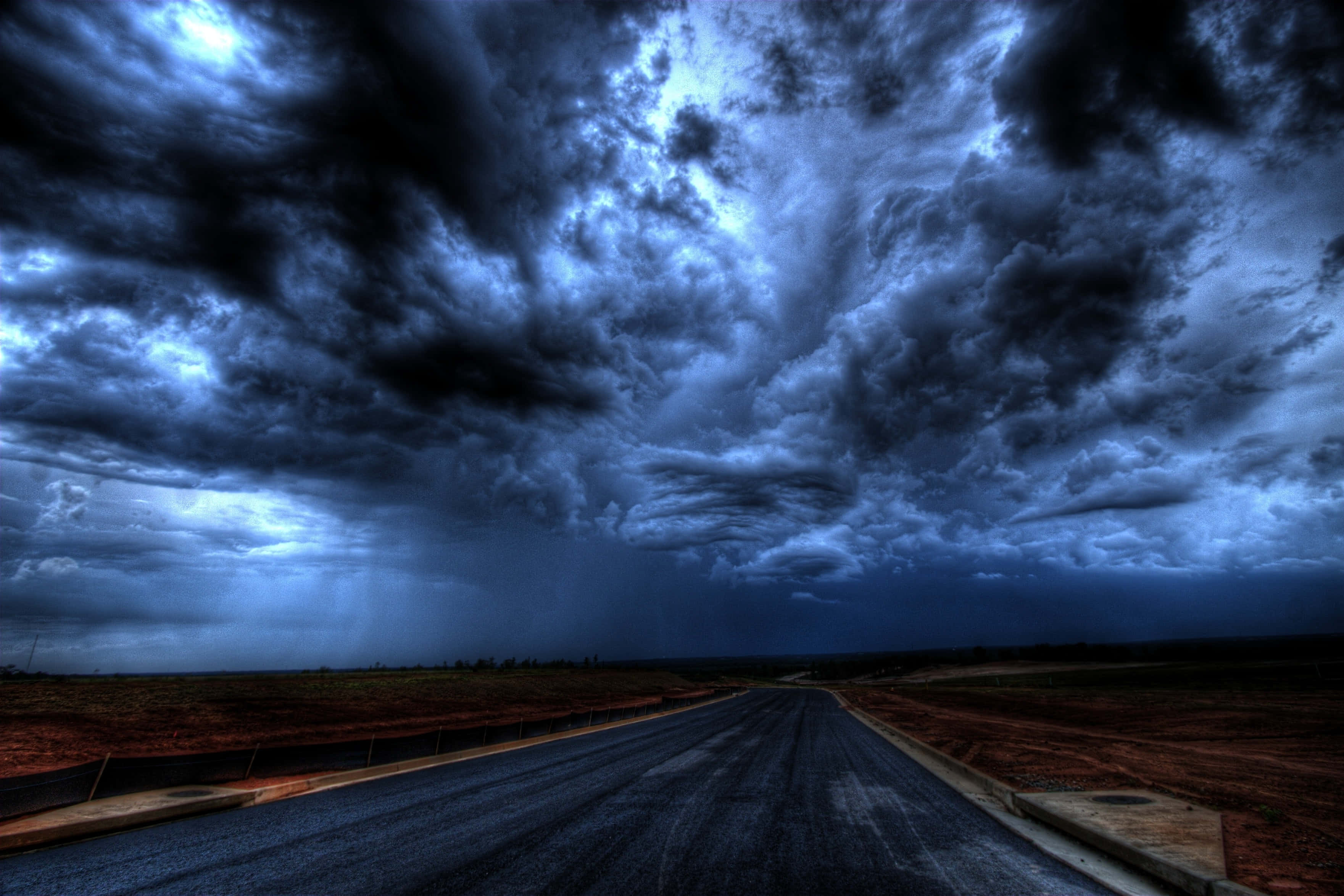 Dark Sky 桌布 壁紙 — 一條孤獨的瀝青路延伸至暴風雨的地平線，天空佈滿戲劇性的藍色和黑色雲層。