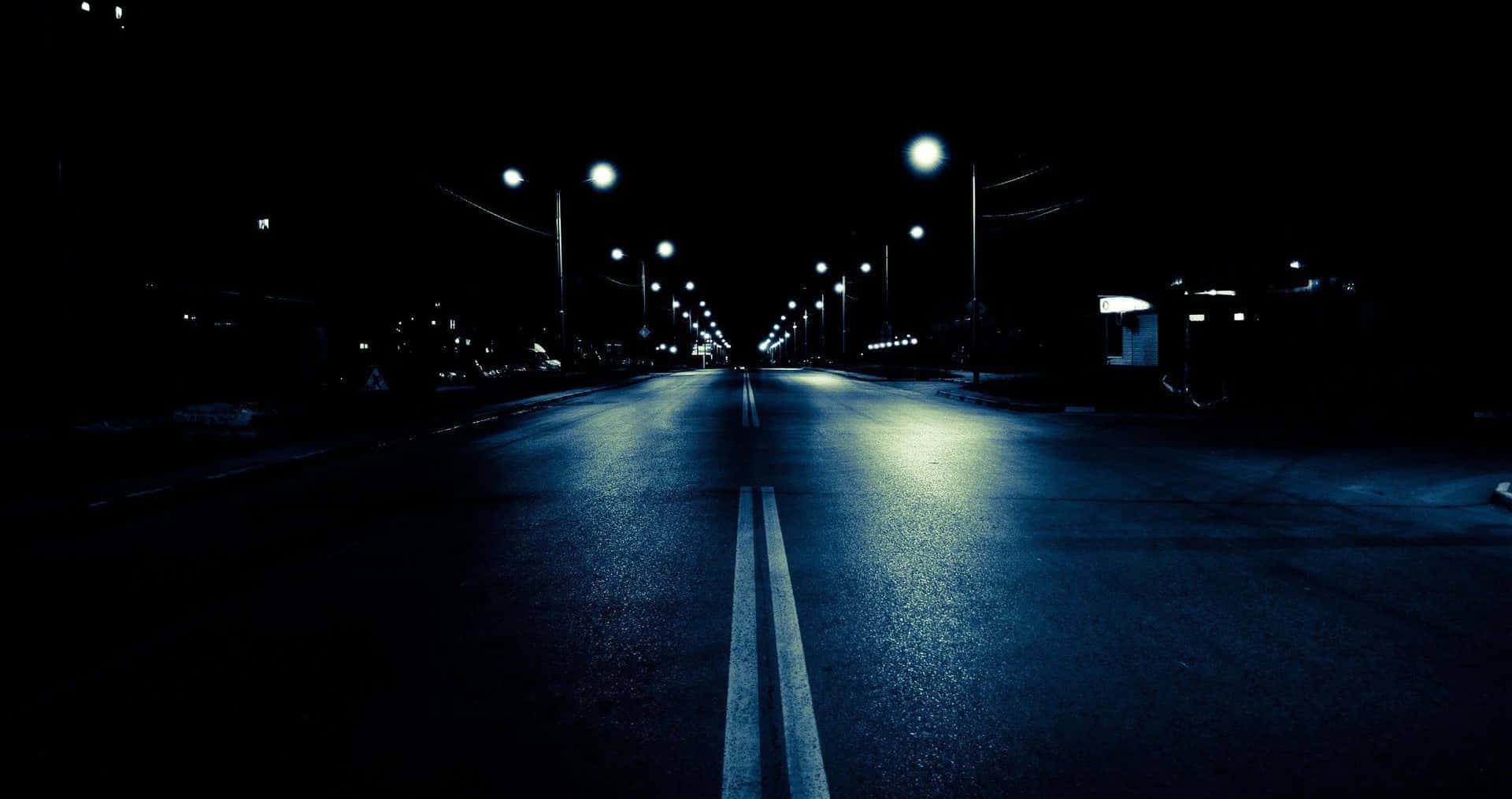 Dark Crossroad 桌布 壁紙 — 一條孤獨的空蕩道路在發光街燈下延伸入黑暗，燈光在濕瀝青上投下藍色倒影。