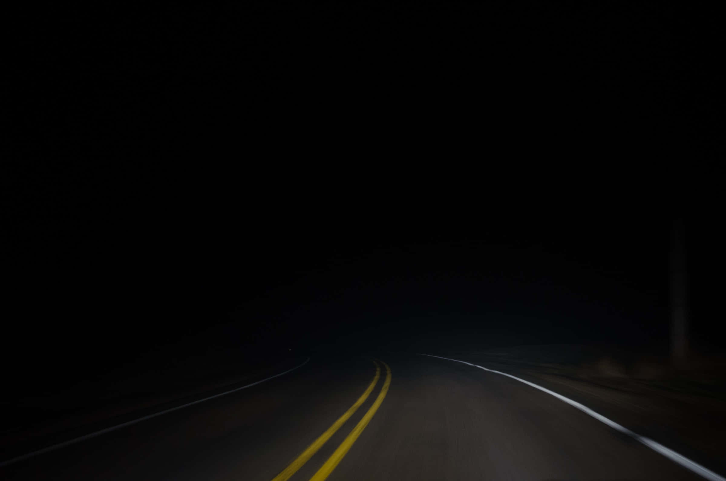 Dark Road 桌布 壁紙 — 一條孤獨的道路在夜間彎入黑暗，僅由微弱的黃色和白色車道標線照亮。