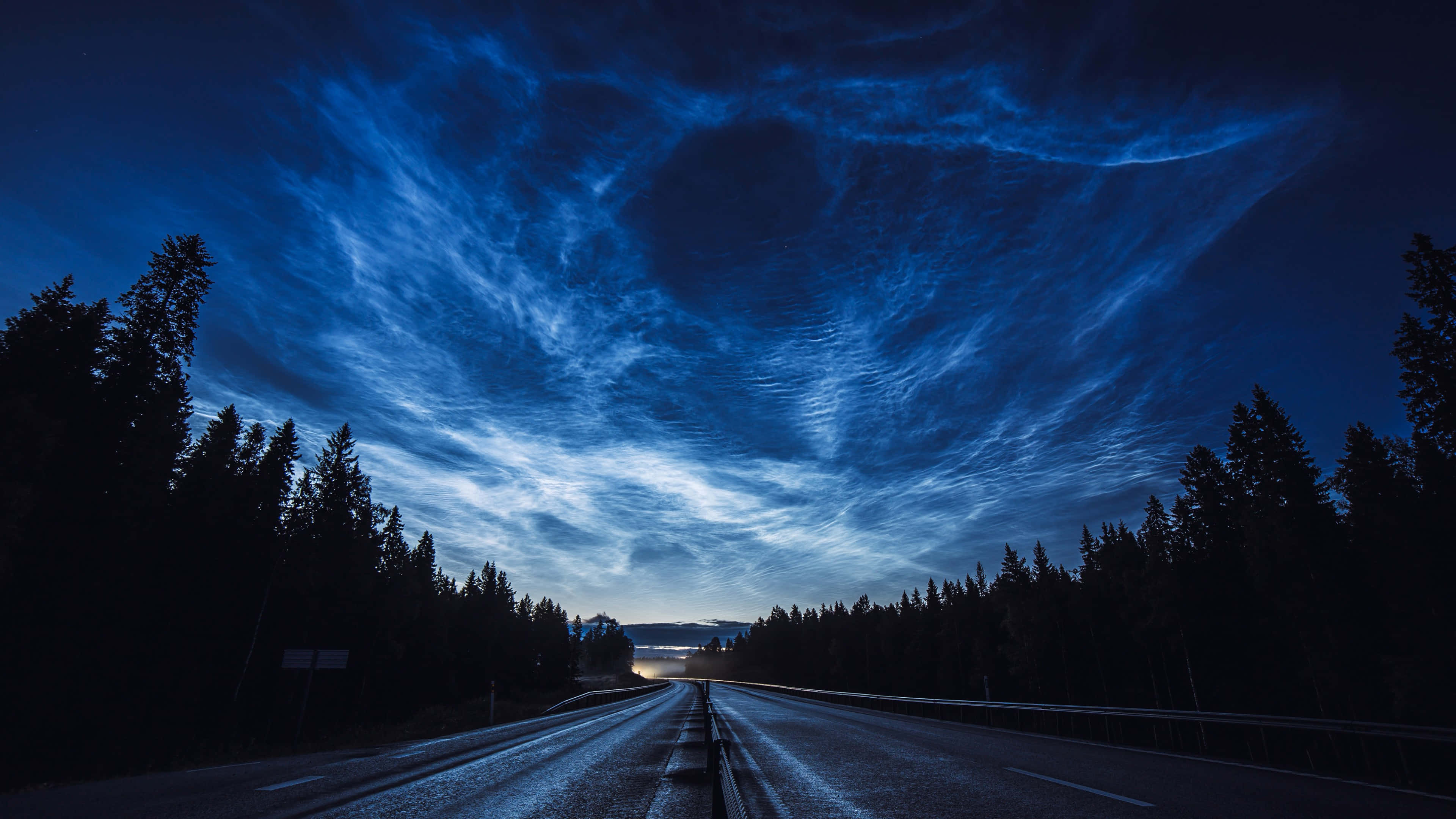 Night Forest 桌布 壁紙 — 一條孤獨的道路在戲劇性的天空下延伸入暮色，天空中有旋轉的藍色夜光雲，兩旁是黑暗的松樹林。