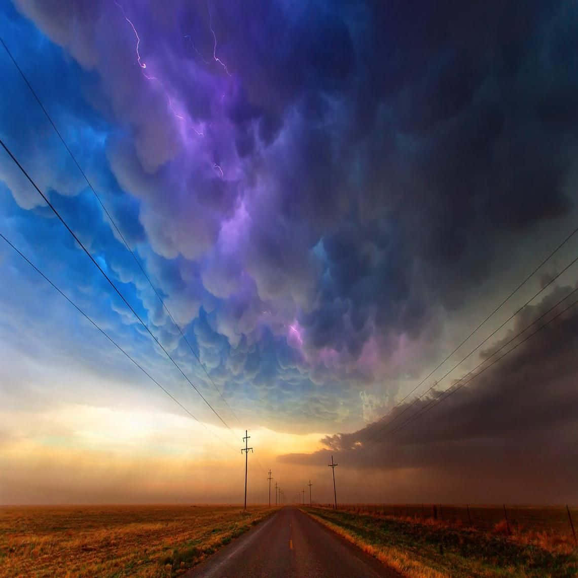 Colorful Sky 桌布 壁紙 — 一條孤獨的道路延伸向戲劇性的天空，紫色閃電在日落時分穿過高聳的風暴雲層。