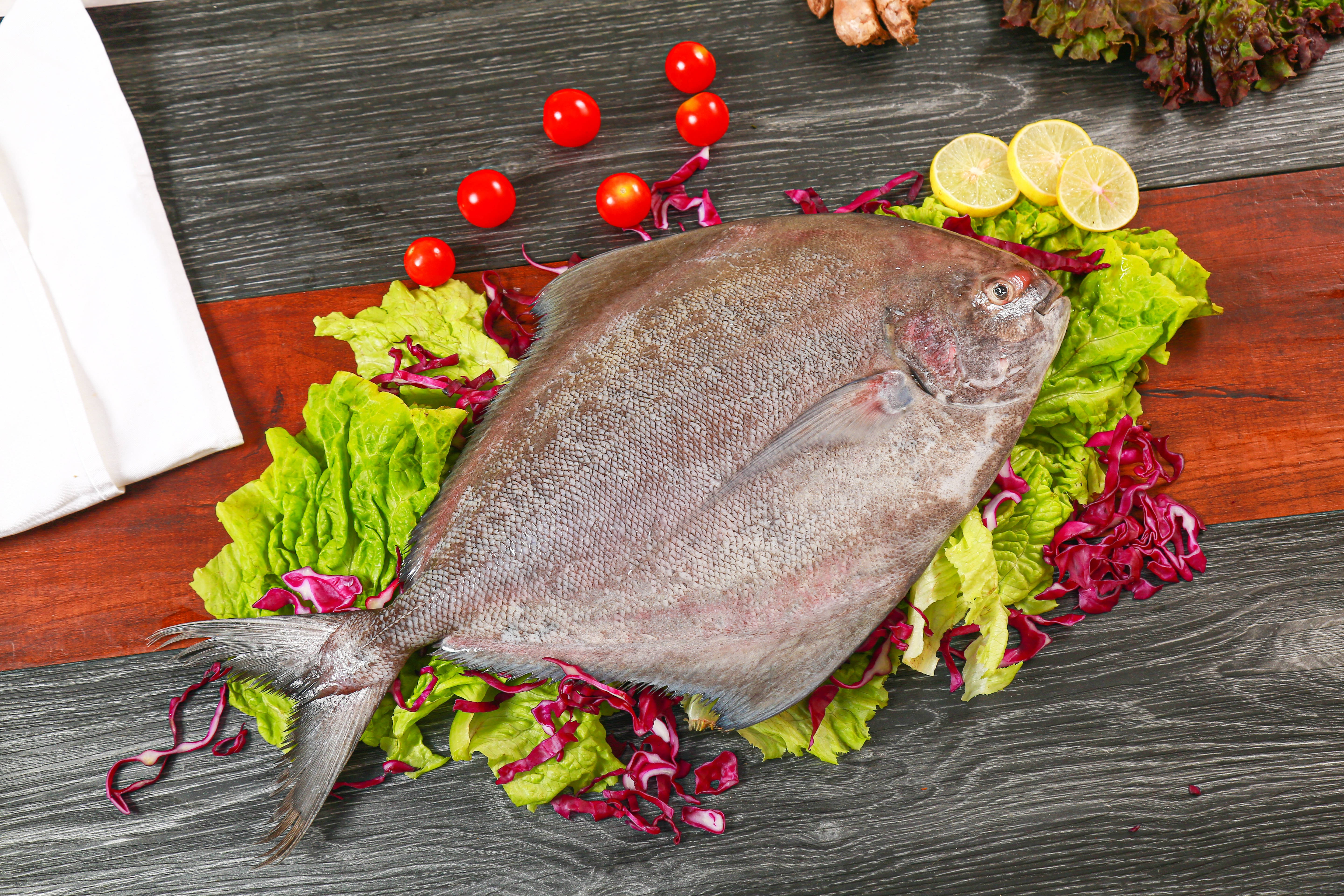 魚 桌布 壁紙 — 一條完整的銀色鰈魚躺在綠色和紫色生菜床上，配上櫻桃番茄和檸檬片，放在深色木質表面上。