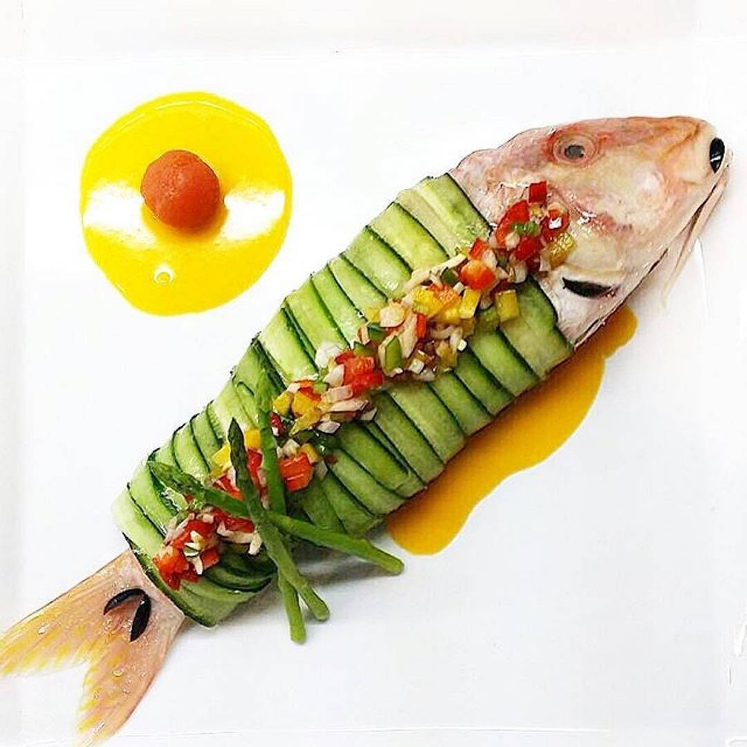 Zucchini 桌布 壁紙 — 一條完整的魚優雅地包裹在黃瓜片中，上面鋪滿色彩繽紛的切丁蔬菜，放在白色盤子上。