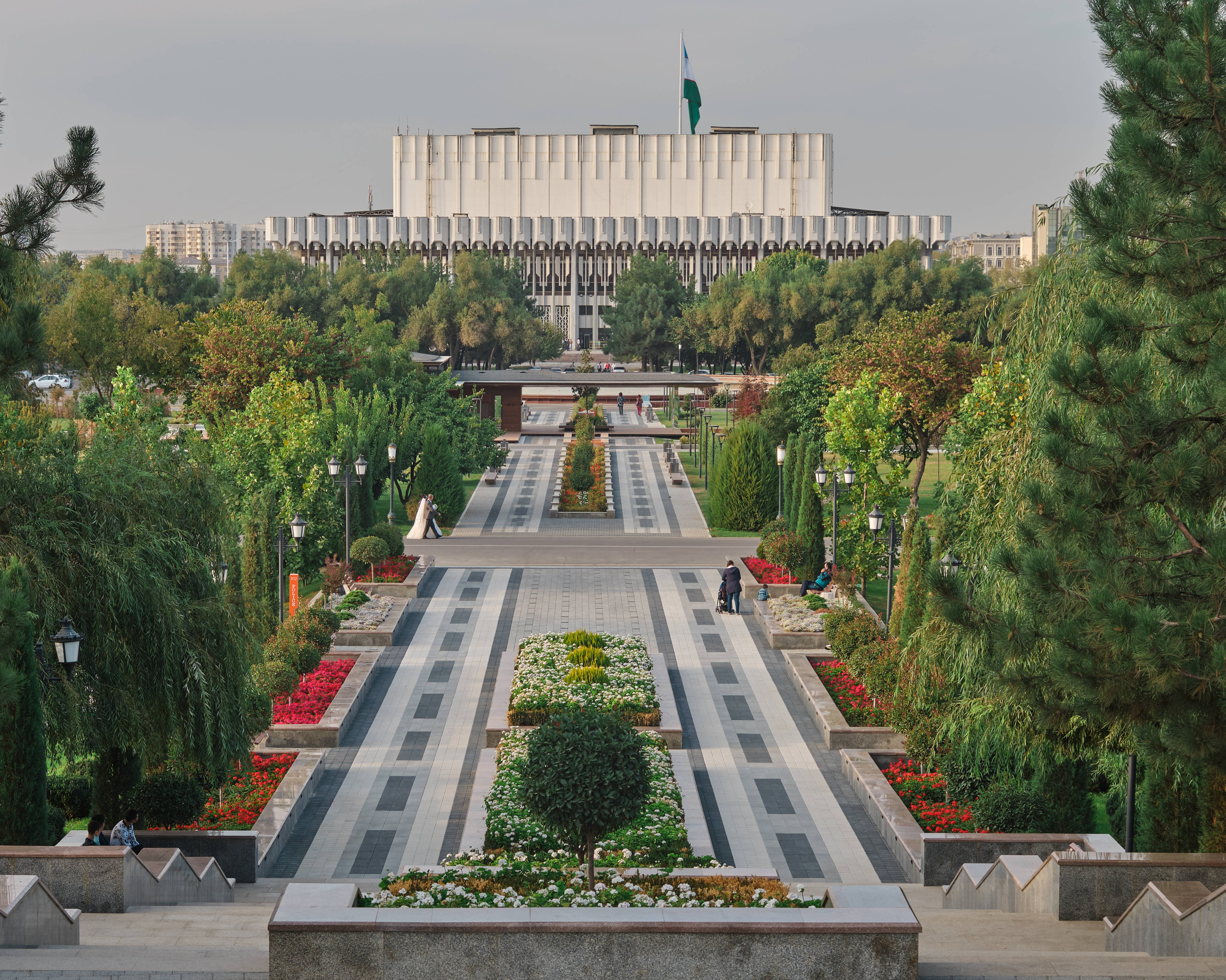 Uzbekistan 桌布 壁紙 — 一條宏偉的對稱花園小徑，兩旁種滿樹木和花壇，通向一座陰天下的現代化大型政府大樓。