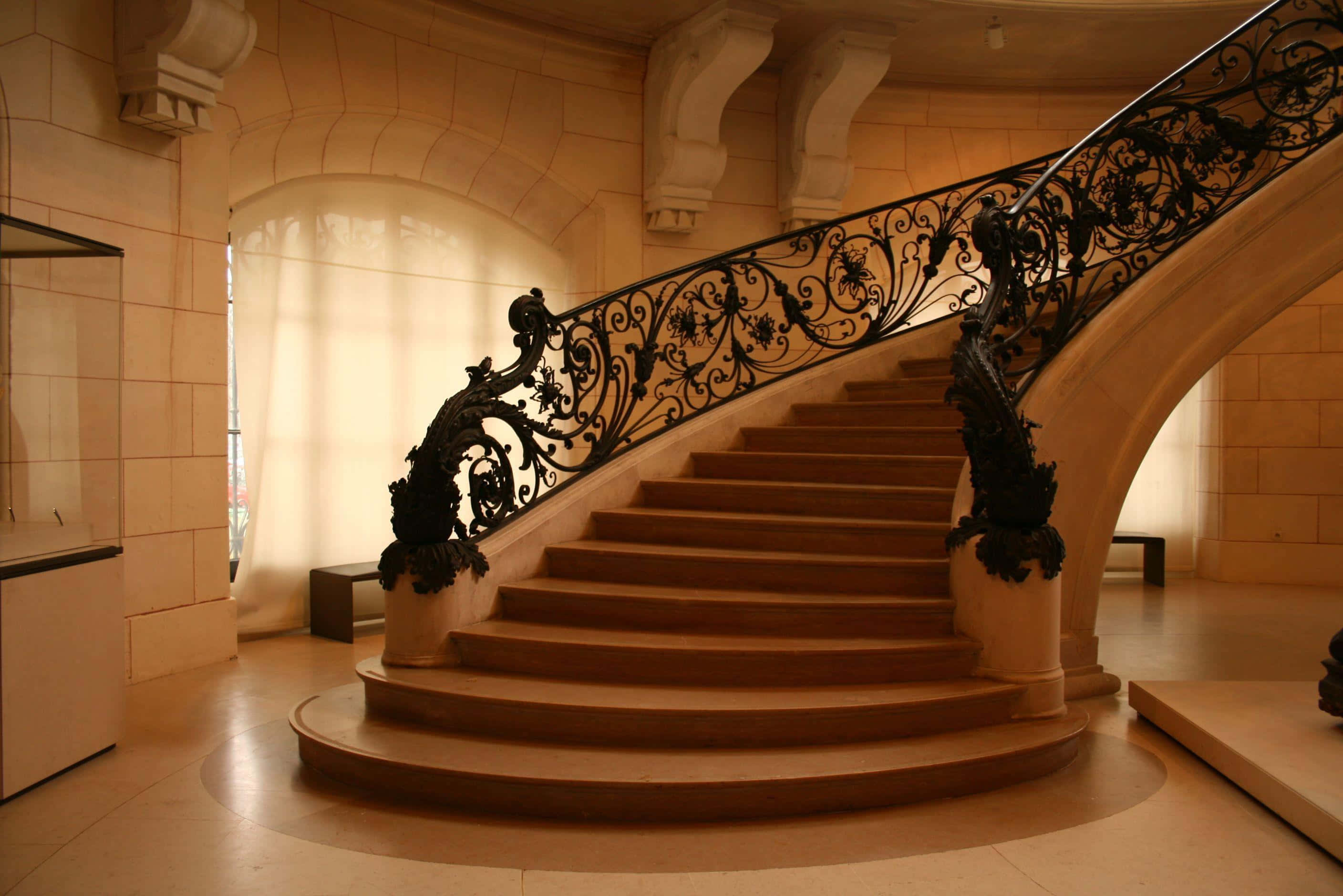 stairs 桌布 壁紙 — 一條宏偉的弧形樓梯，帶有華麗的黑色鐵欄杆，穿過一個溫暖照明的石廳，拱形窗戶。