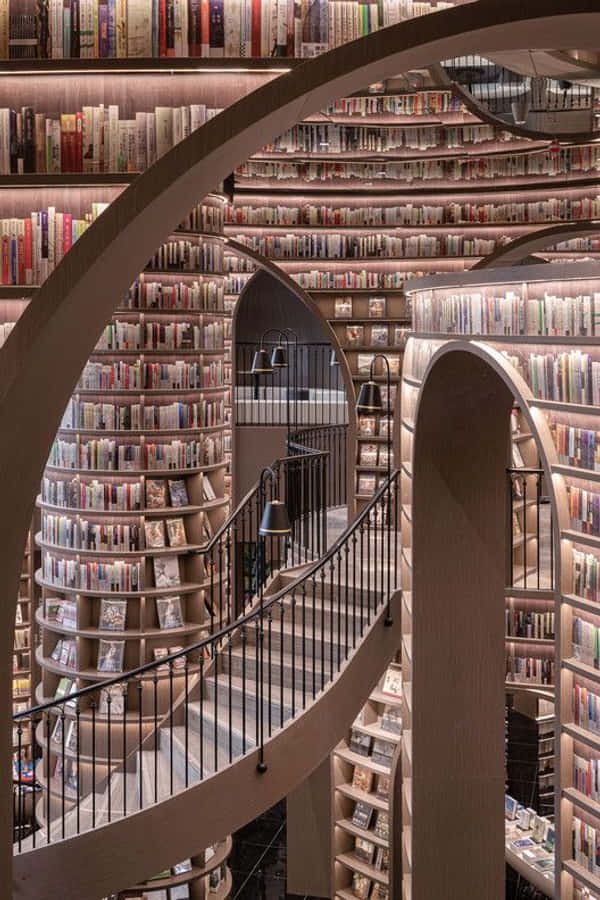 Library 桌布 壁紙 — 一條宏偉的螺旋樓梯蜿蜒穿過高聳的木製書架，書架上擺滿了書籍，位於一個燈光溫暖的現代圖書館內。