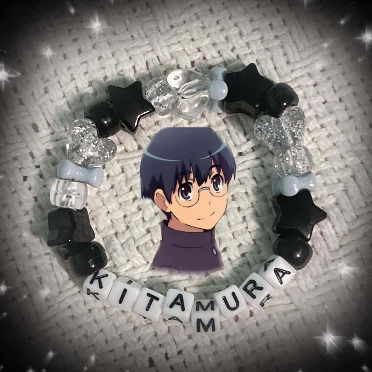 Yusaku Kitamura 桌布 壁紙 — 一條客製化珠串手鍊，用黑色星星和亮粉珠子在紋理白色布料背景上拼出Kitamura，圍繞一個印刷的動漫角色肖像。