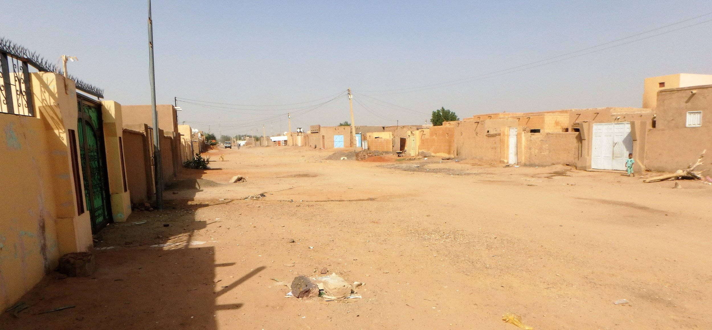 Sudan 桌布 壁紙 — 一條寧靜塵土飛揚的街道，位於陽光普照的村莊，有土坯建築和稀疏的植被，在清澈的藍天下。