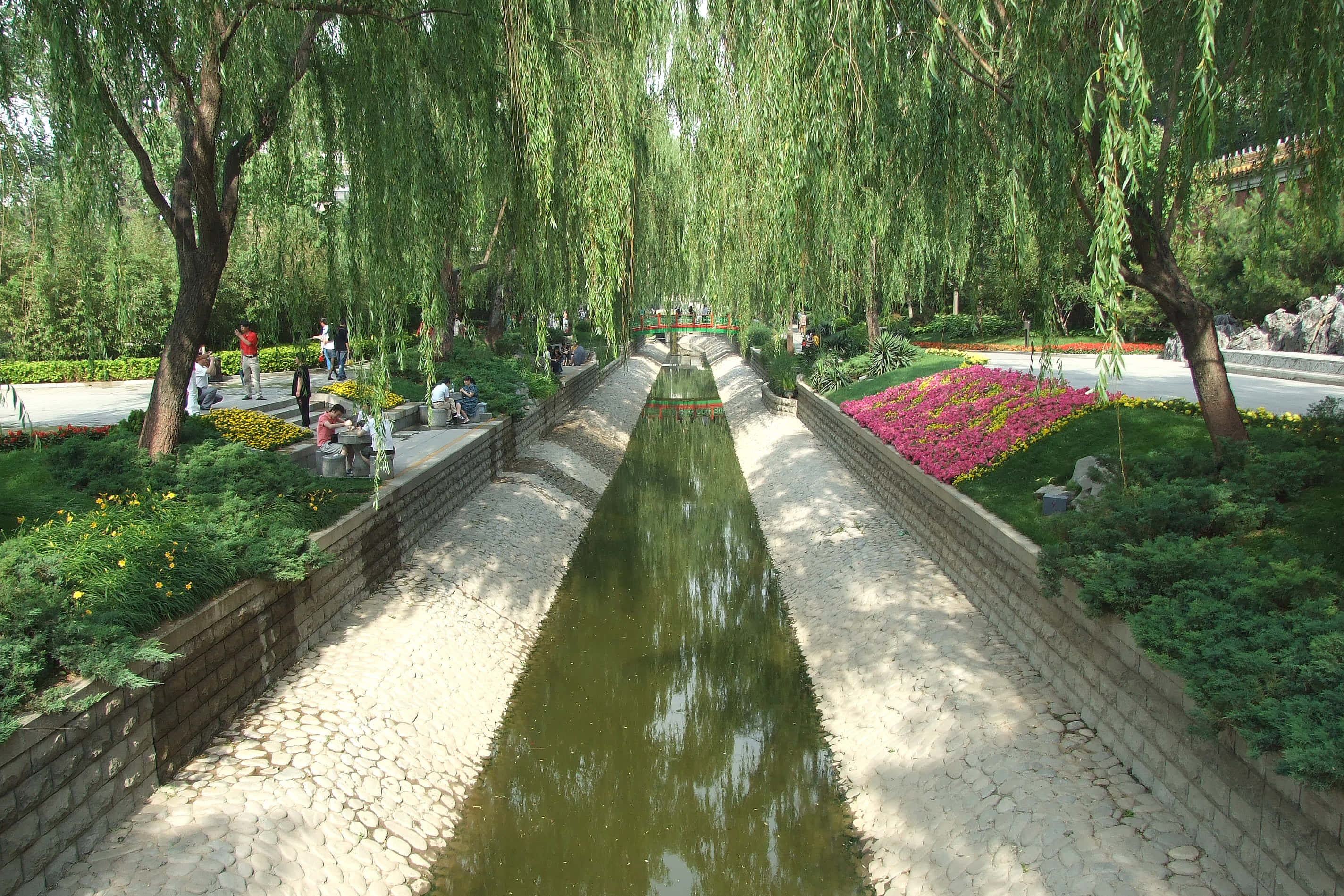 公園 桌布 壁紙 — 一條寧靜的公園小徑，兩旁種滿垂柳和色彩繽紛的花壇，人們在平靜的水道旁放鬆身心。