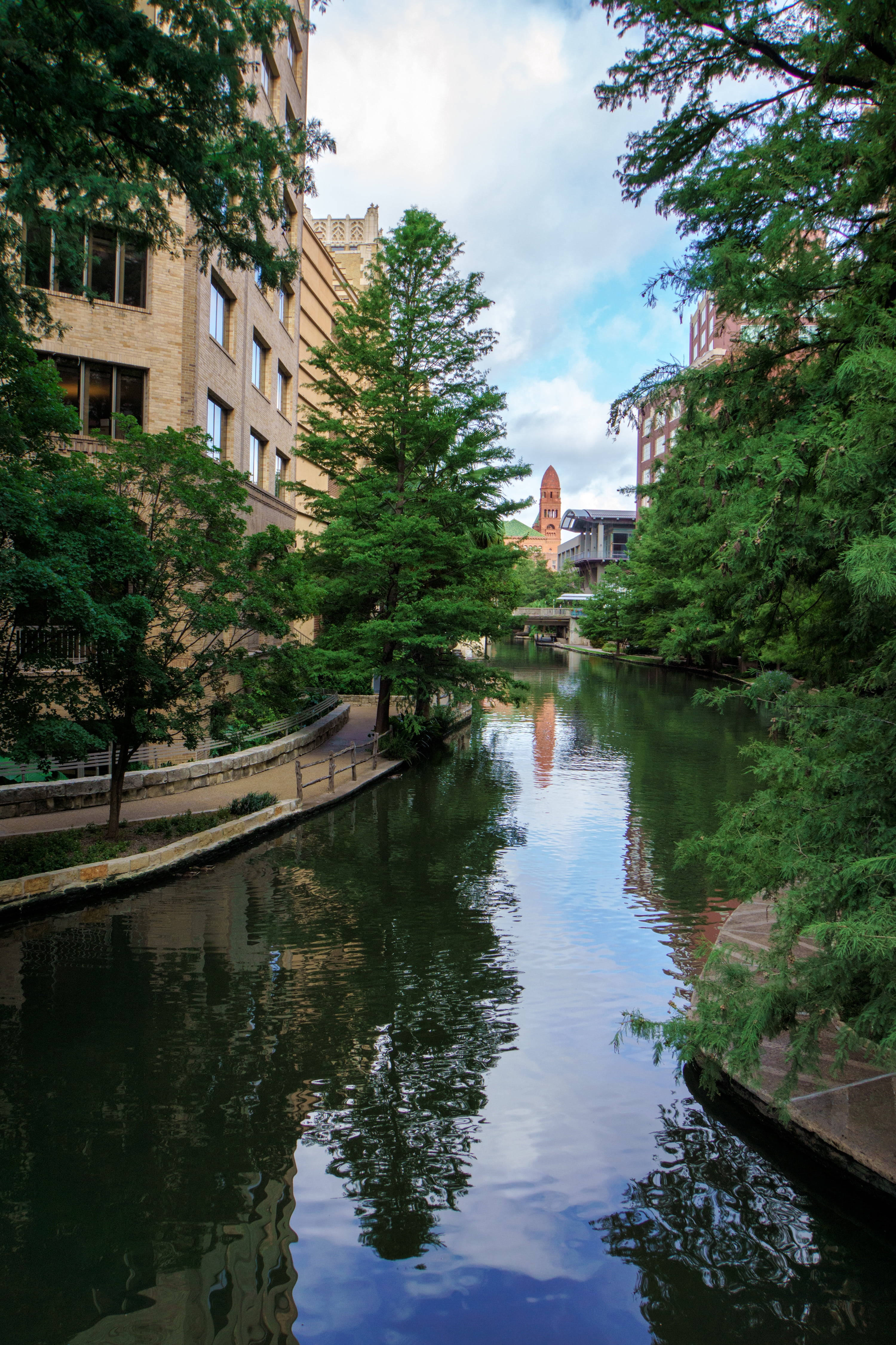 San Antonio 桌布 壁紙 — 一條寧靜的城市運河，兩旁種滿茂密的綠樹和歷史悠久的磚砌建築，天空部分多雲。