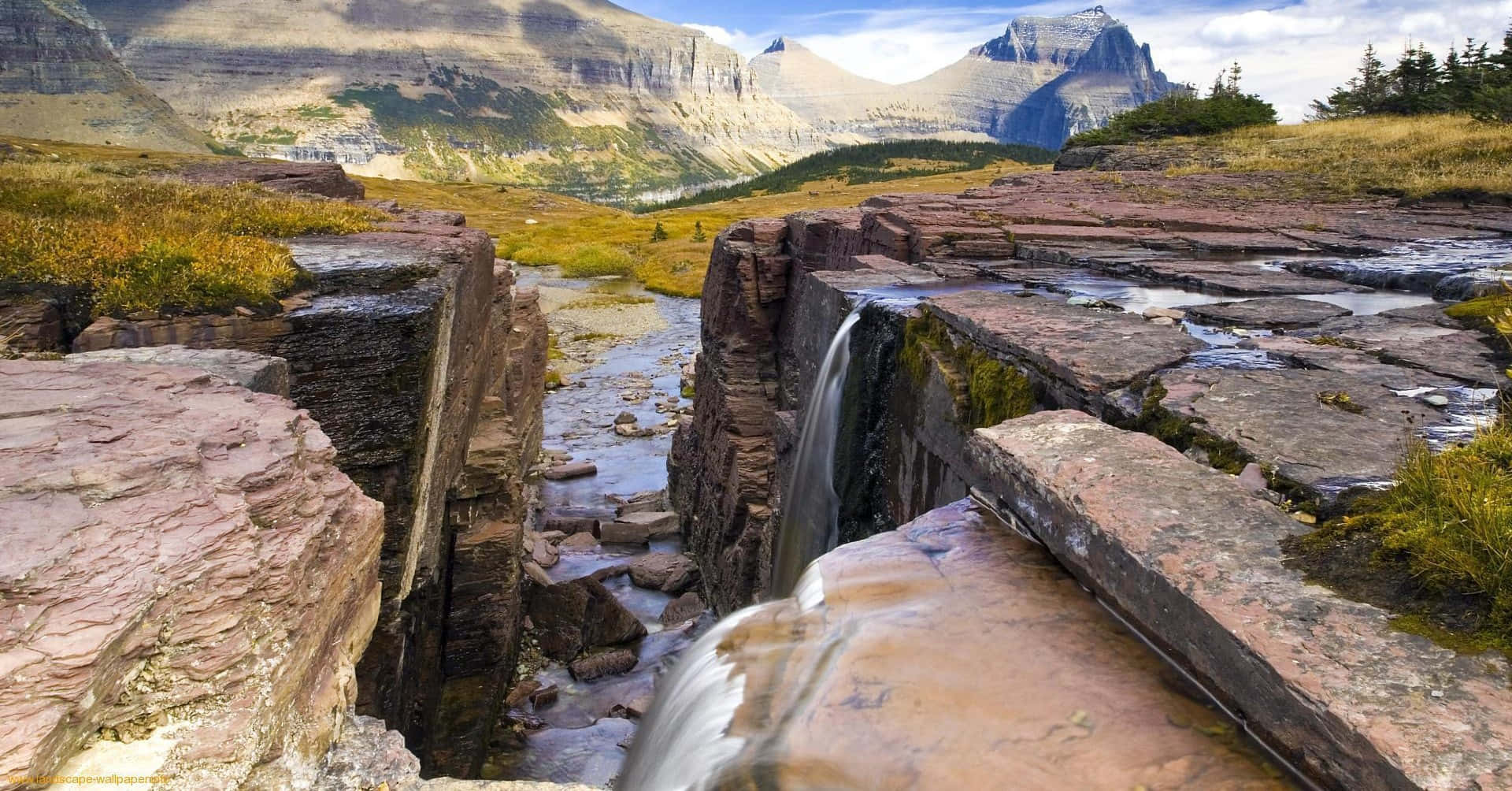 Montana 桌布 壁紙 — 一條寧靜的山間溪流，流過層層疊疊的粉棕色岩石，穿過崎嶇的山谷，天空局部多雲。