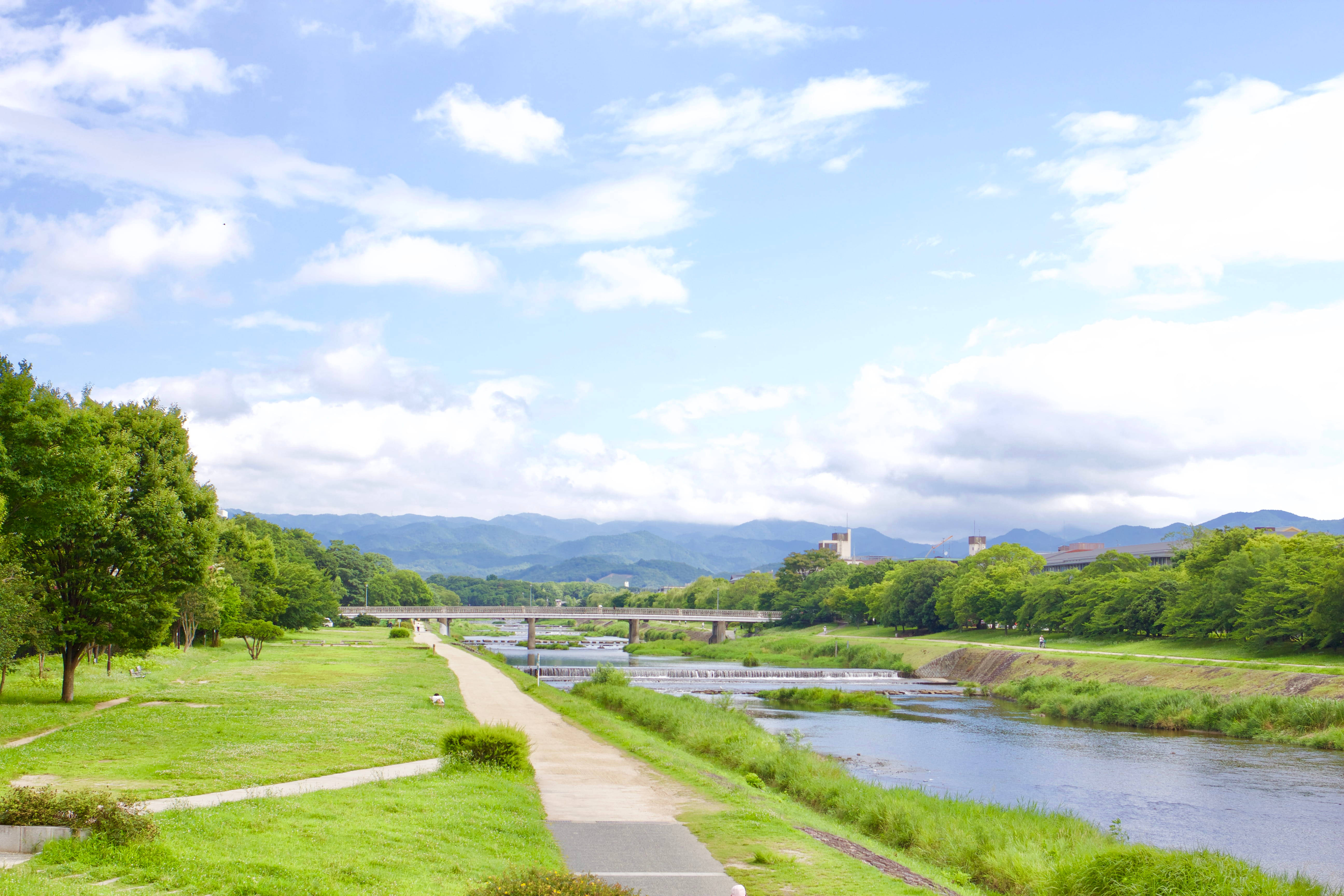 Road 桌布 壁紙 — 一條寧靜的河流流經一個綠意盎然的公園，有蜿蜒的小徑和遠處的山脈，在明亮的藍天下有蓬鬆的雲朵。