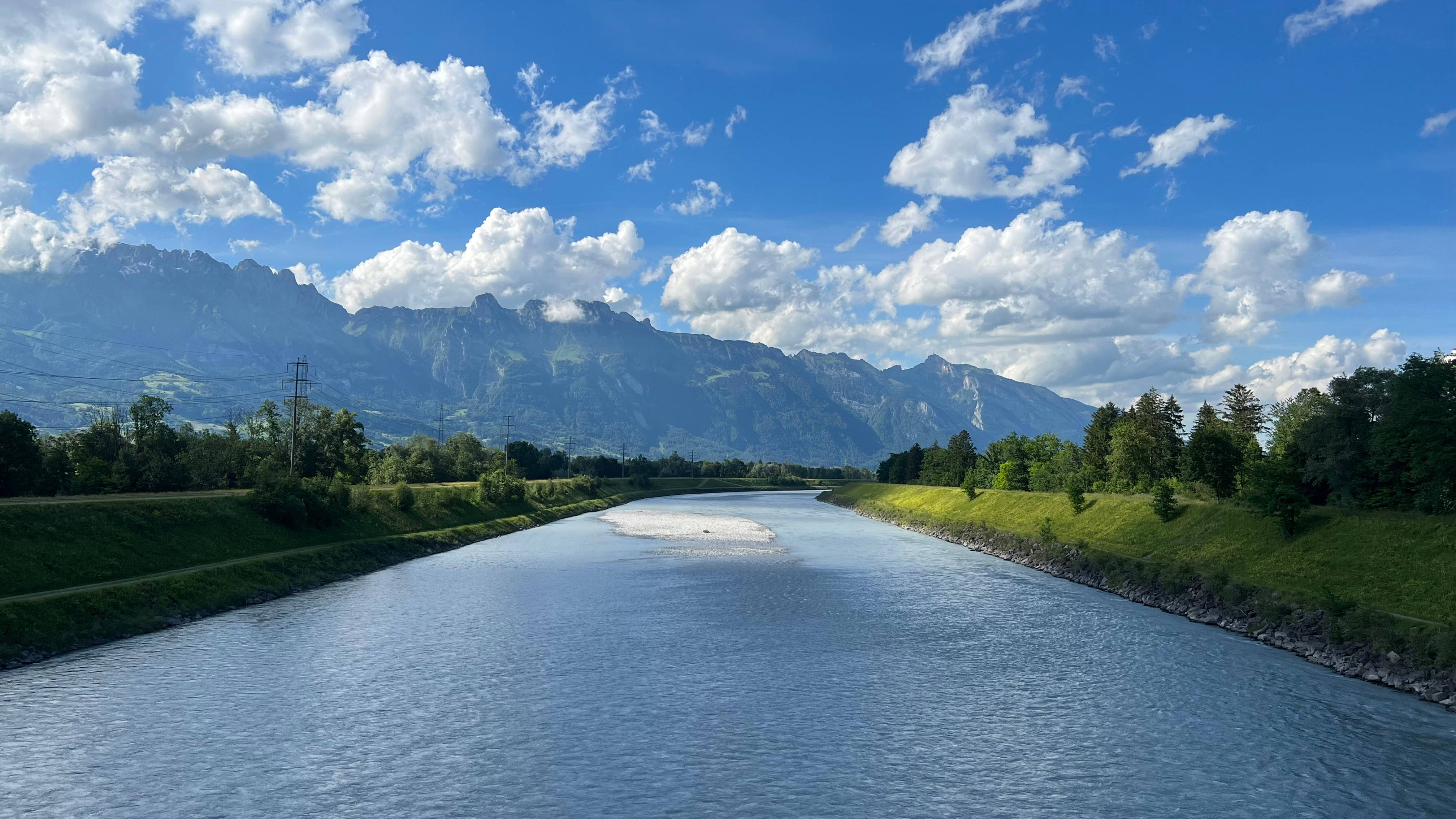 Liechtenstein 桌布 壁紙 — 一條寧靜的河流流經鬱鬱蔥蔥的綠色河岸，上方是明亮的藍天，點綴著蓬鬆的白雲，遠處有雄偉的山脈。